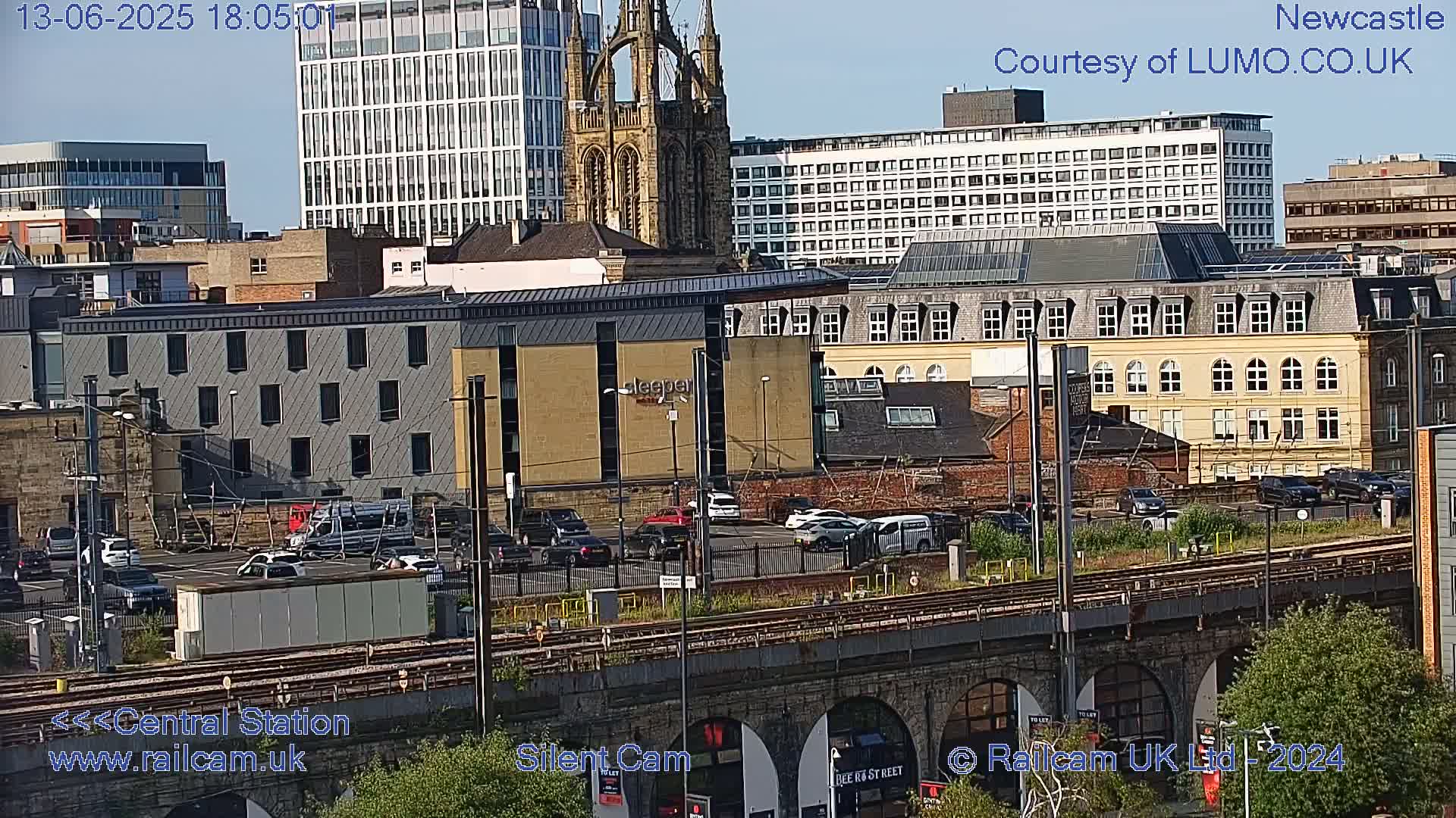 A high-angle, long shot shows a city landscape on a sunny day, featuring various buildings, parked cars, train tracks, and some greenery.