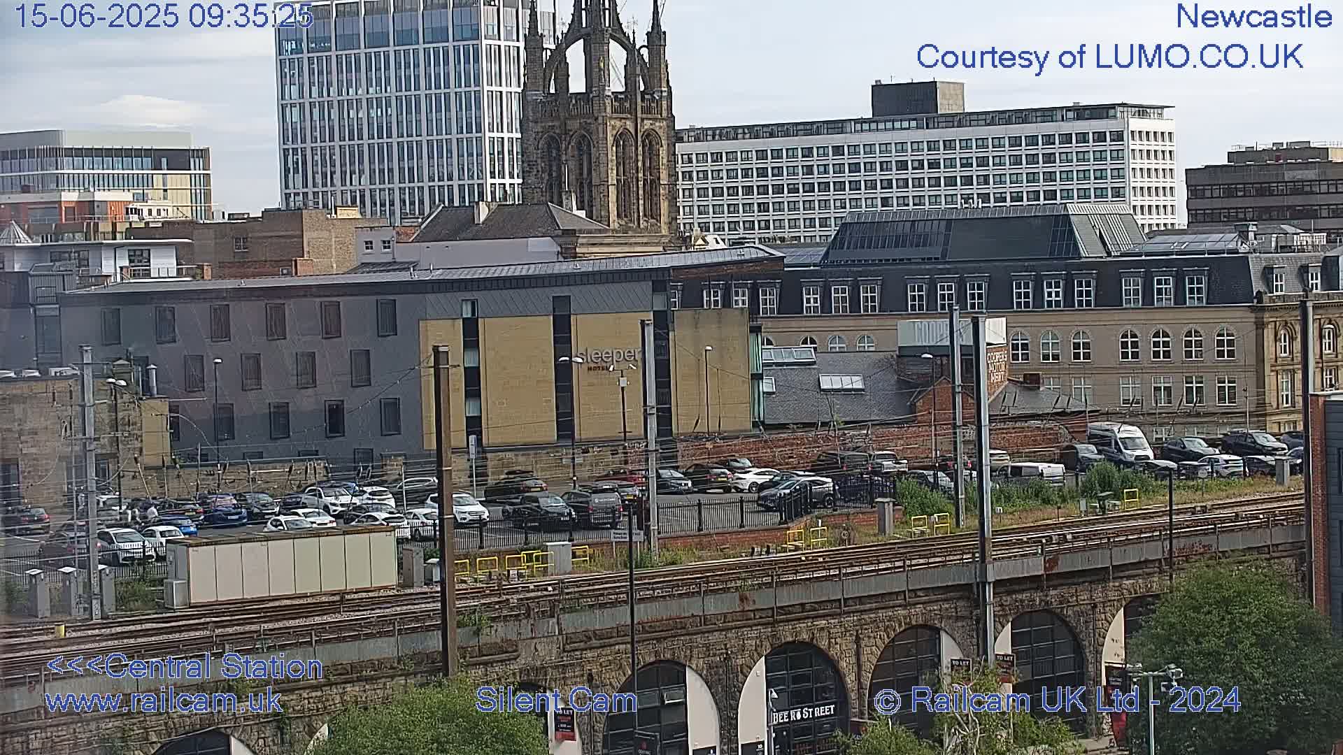 A city view of a train station, numerous buildings, and a parking lot on a partly cloudy day.