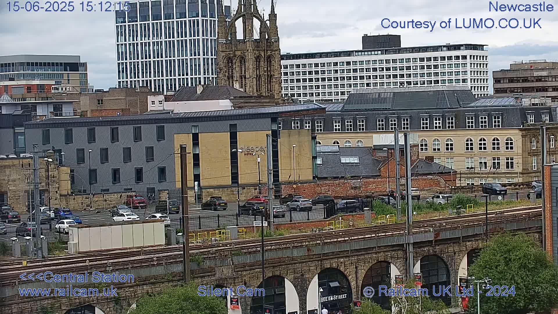 A city scene under an overcast sky shows a parking lot, train tracks, and various buildings including a church and modern high-rises.