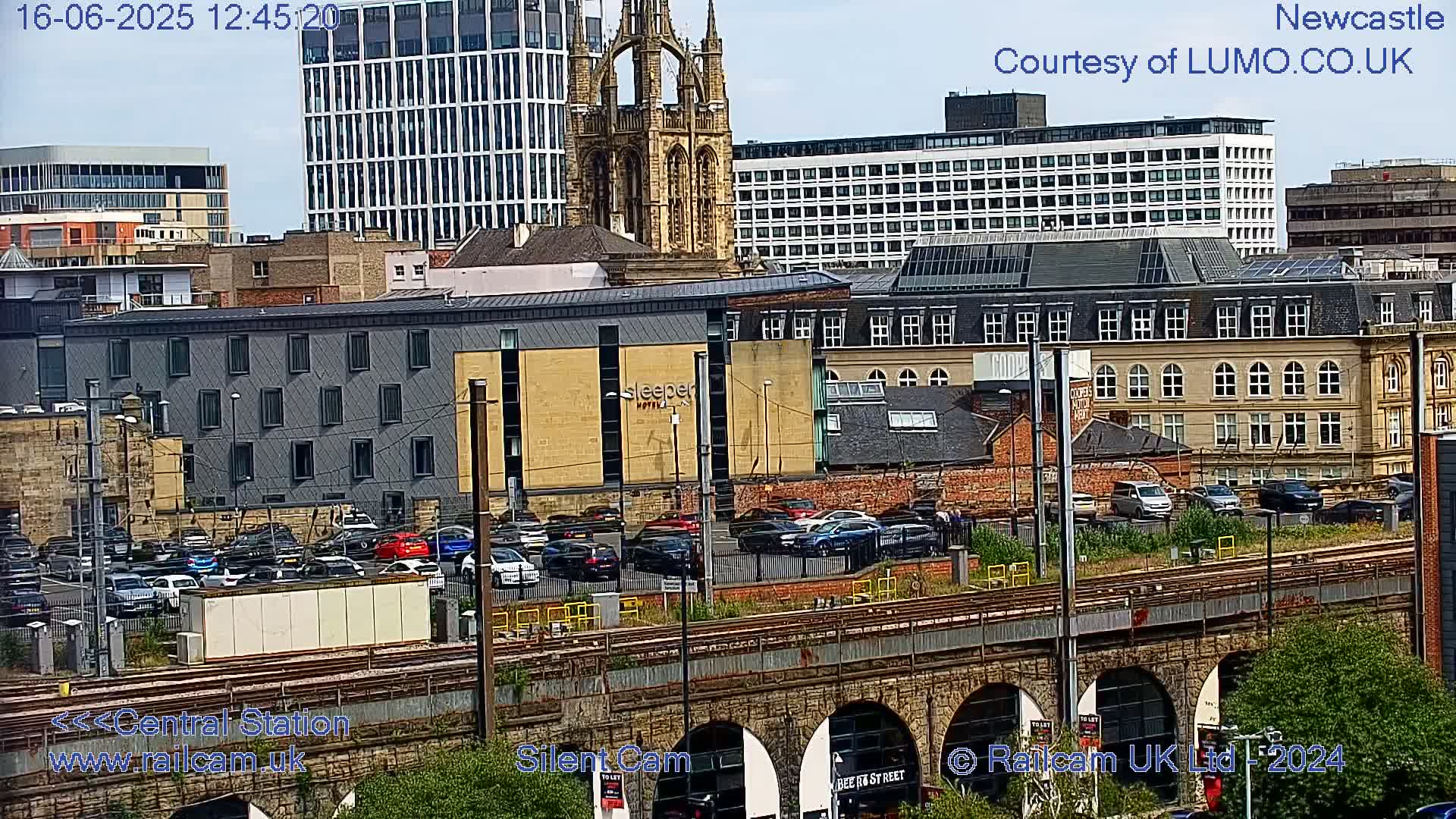 A city scene on a partly cloudy day shows a variety of buildings, including a church, modern high-rises, and several older structures, with a parking lot and train tracks in the foreground.