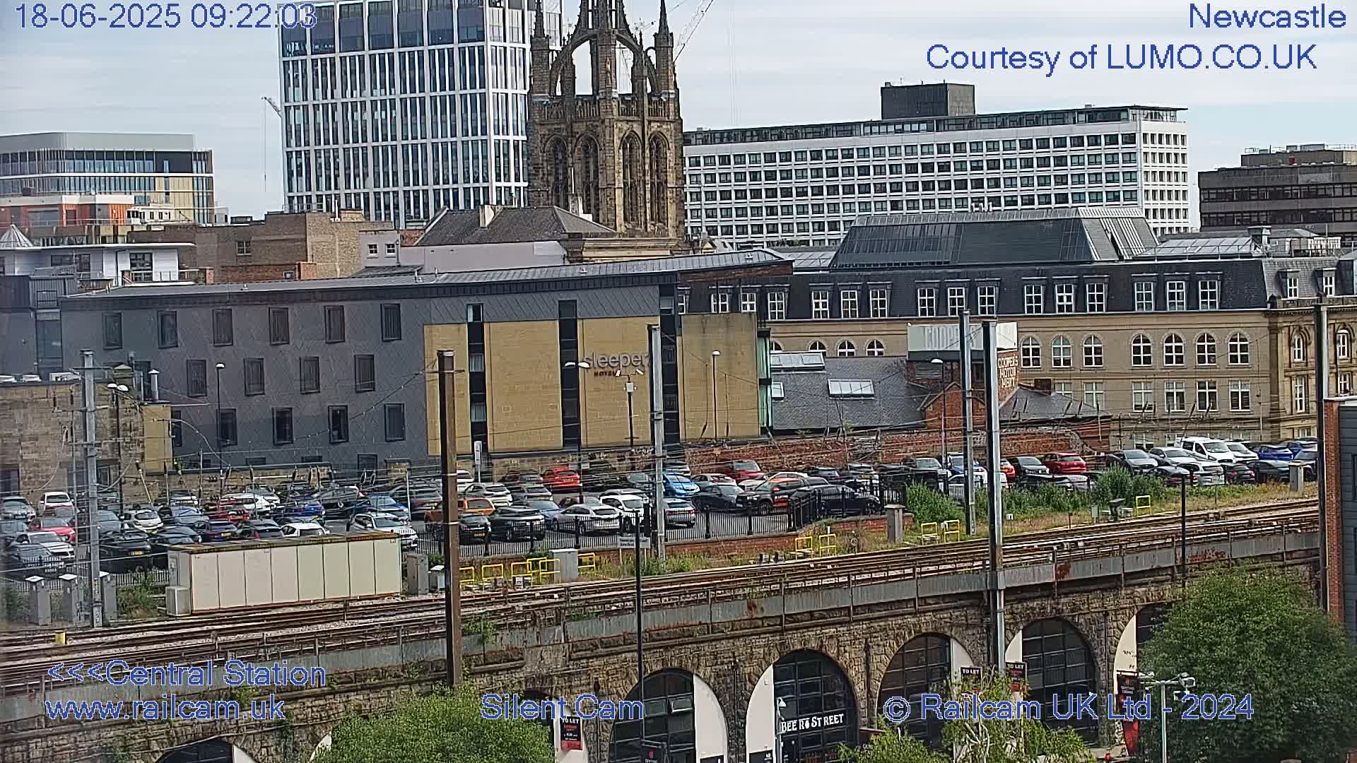 A city landscape with a parking lot full of cars, train tracks, and various buildings including a tall modern building and an older stone church, under an overcast sky.