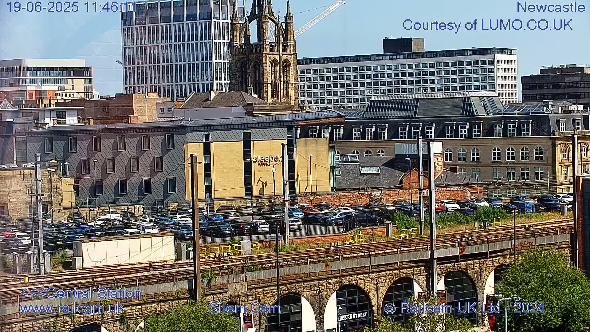 A sunny view of a city with numerous buildings, a parking lot full of cars, and railway tracks spanning an arched stone structure.