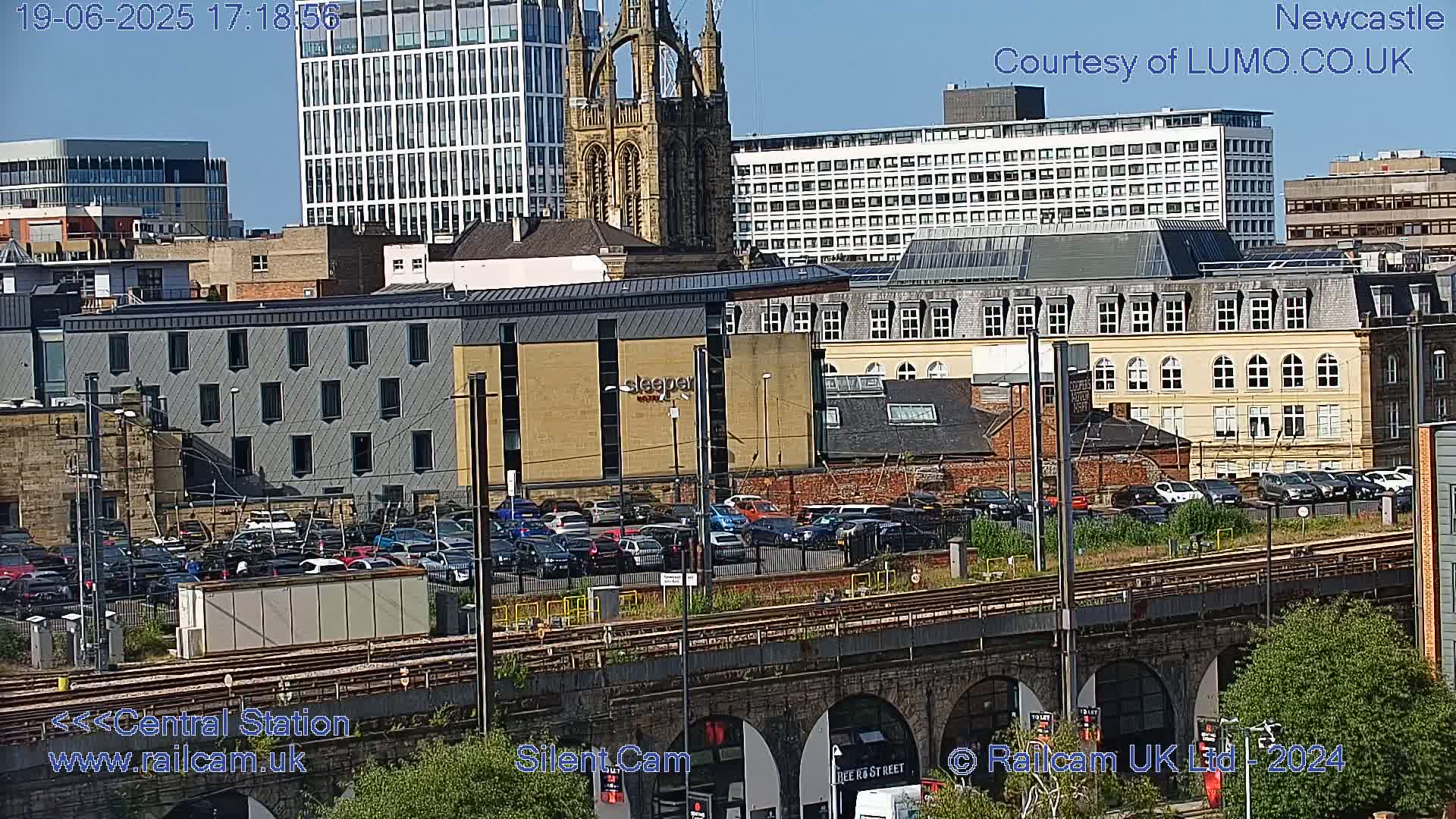 A city scene on a sunny day shows a large parking lot filled with cars next to train tracks that run under a bridge; various buildings, including a tall modern building and a church spire, are visible in the background.