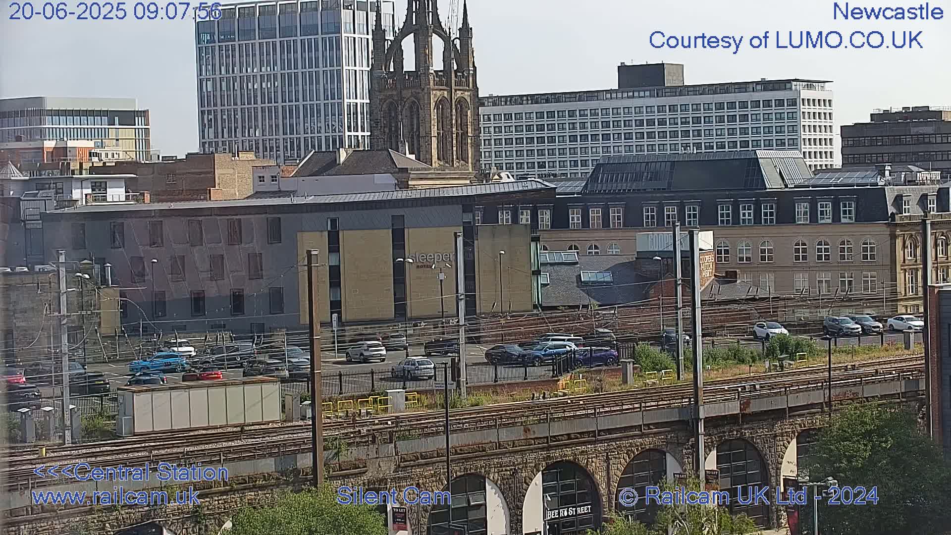 A sunny view of a city skyline featuring several buildings, a church steeple, train tracks, and parked cars.
