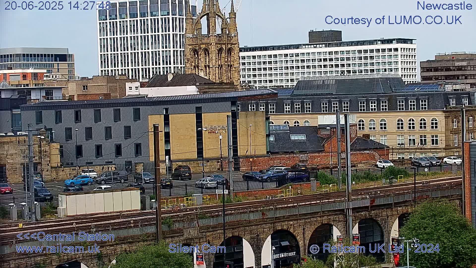A city scene viewed from a distance shows several buildings, including a church, a hotel, and apartment complexes, with cars parked in a lot near train tracks and an arched stone bridge.