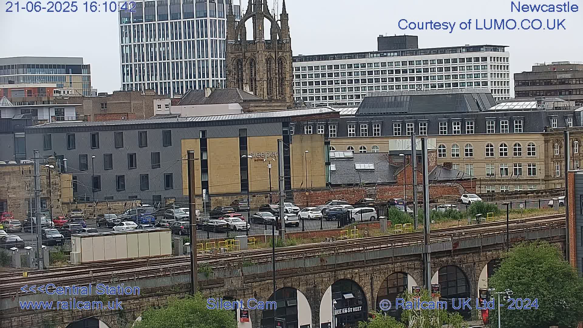 A city view with overcast skies shows a parking lot filled with cars next to a railway track, situated beneath a bridge with stone arches, and various buildings including a tall modern building and a church.
