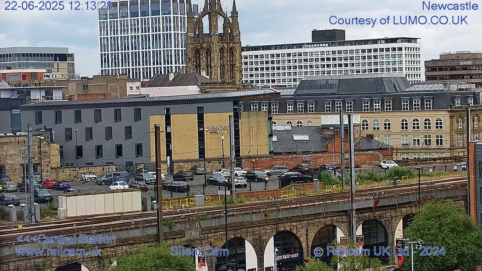 A city scene viewed from above, showing a mix of modern and older buildings, a parking lot filled with cars, and train tracks spanning a stone viaduct, under an overcast sky.