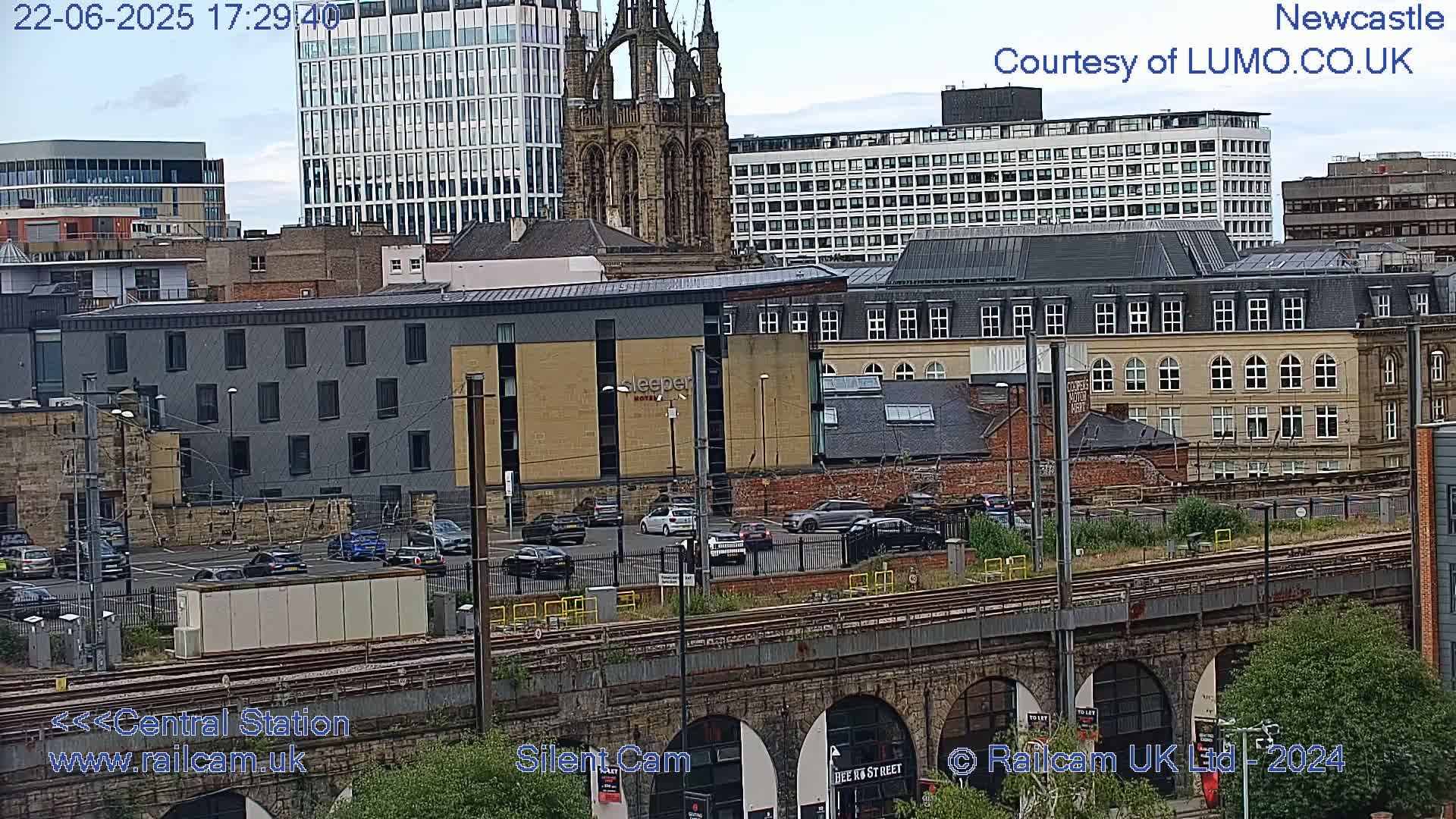 A city scene with overcast skies shows several buildings, including a modern hotel, older brick structures, and a tall church spire, all viewed from above a railway line with cars in a parking lot.