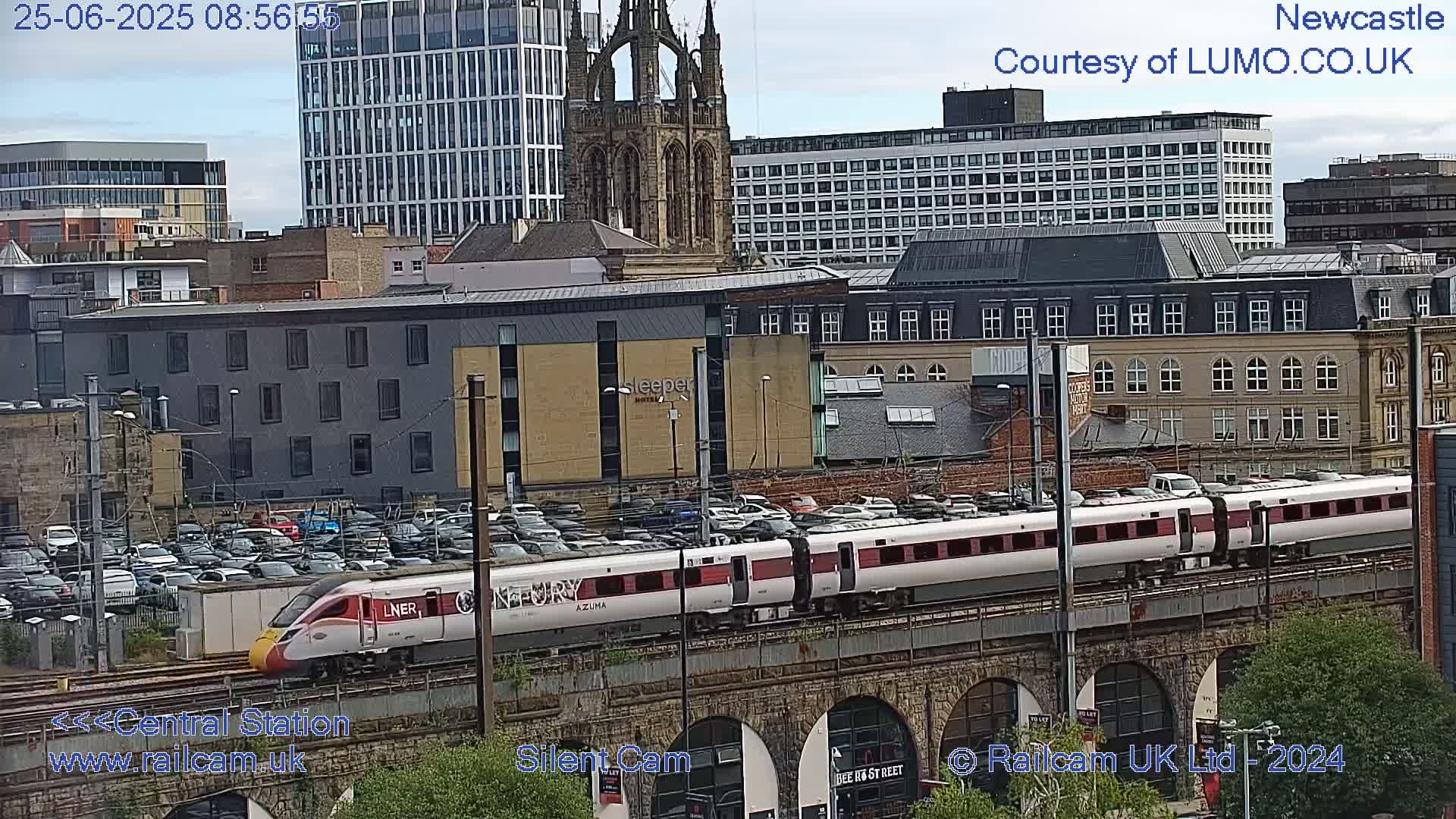 A long white and maroon passenger train passes over a stone viaduct in a city with overcast skies, numerous buildings, and a large parking lot visible.