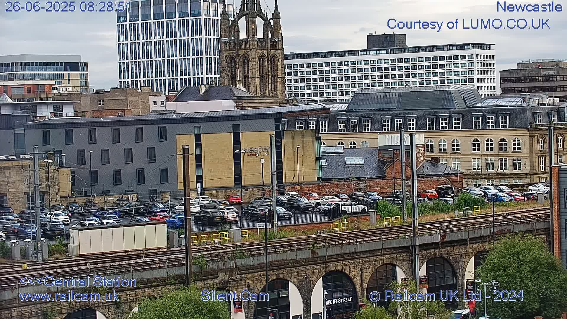 A city view on an overcast day shows a large parking lot filled with cars next to train tracks that curve under an arched stone structure, with various buildings and a tall church spire in the background.