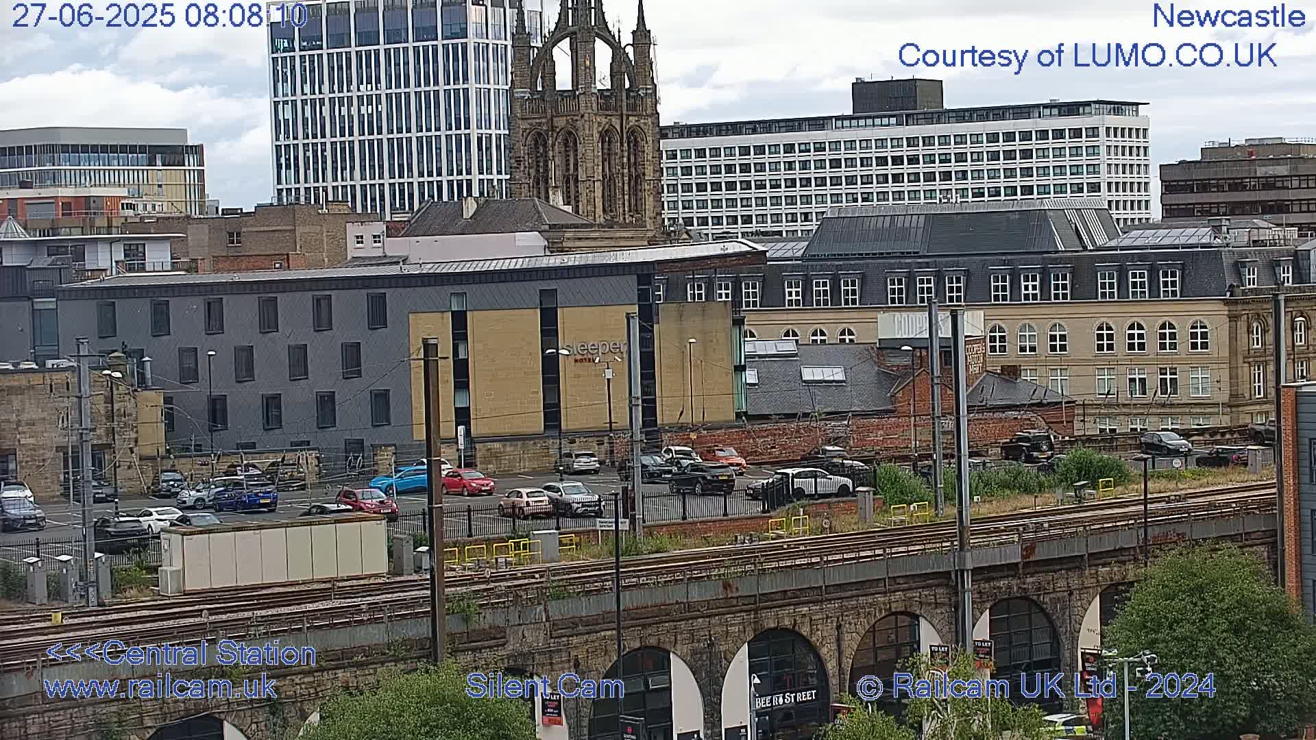 A city scene viewed from above shows a parking lot with cars next to train tracks and various buildings, including a tall modern building and a gothic-style church, under a partly cloudy sky.