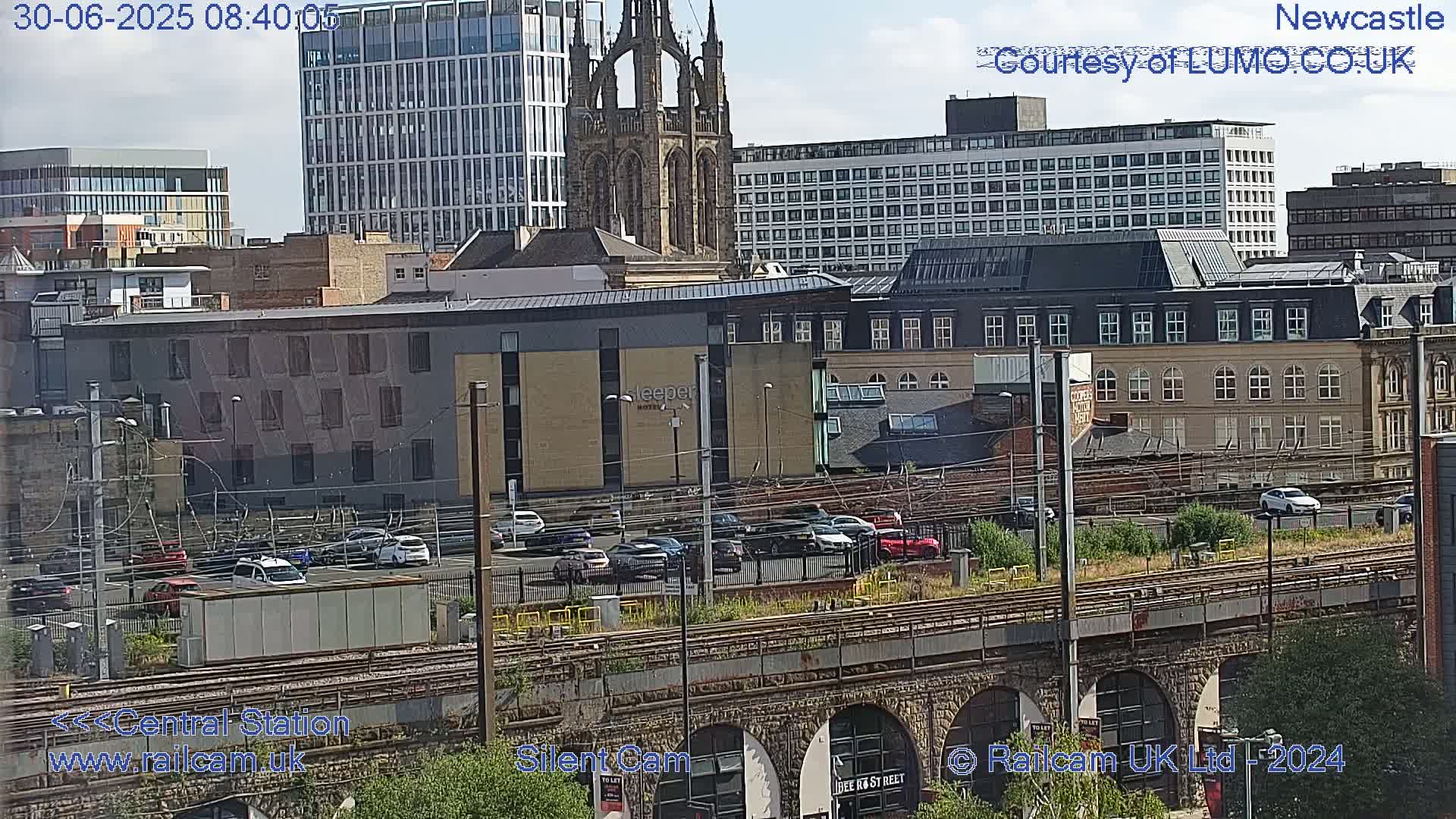 A city skyline under a partly cloudy sky, viewed from above a railway trestle, showing various buildings including a tall church and modern structures, with a parking lot and cars visible below.