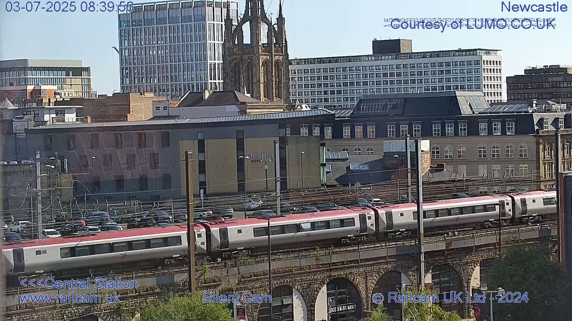 A long passenger train travels across a railway bridge on a sunny day, passing through a city with many buildings of varying heights and architectural styles.