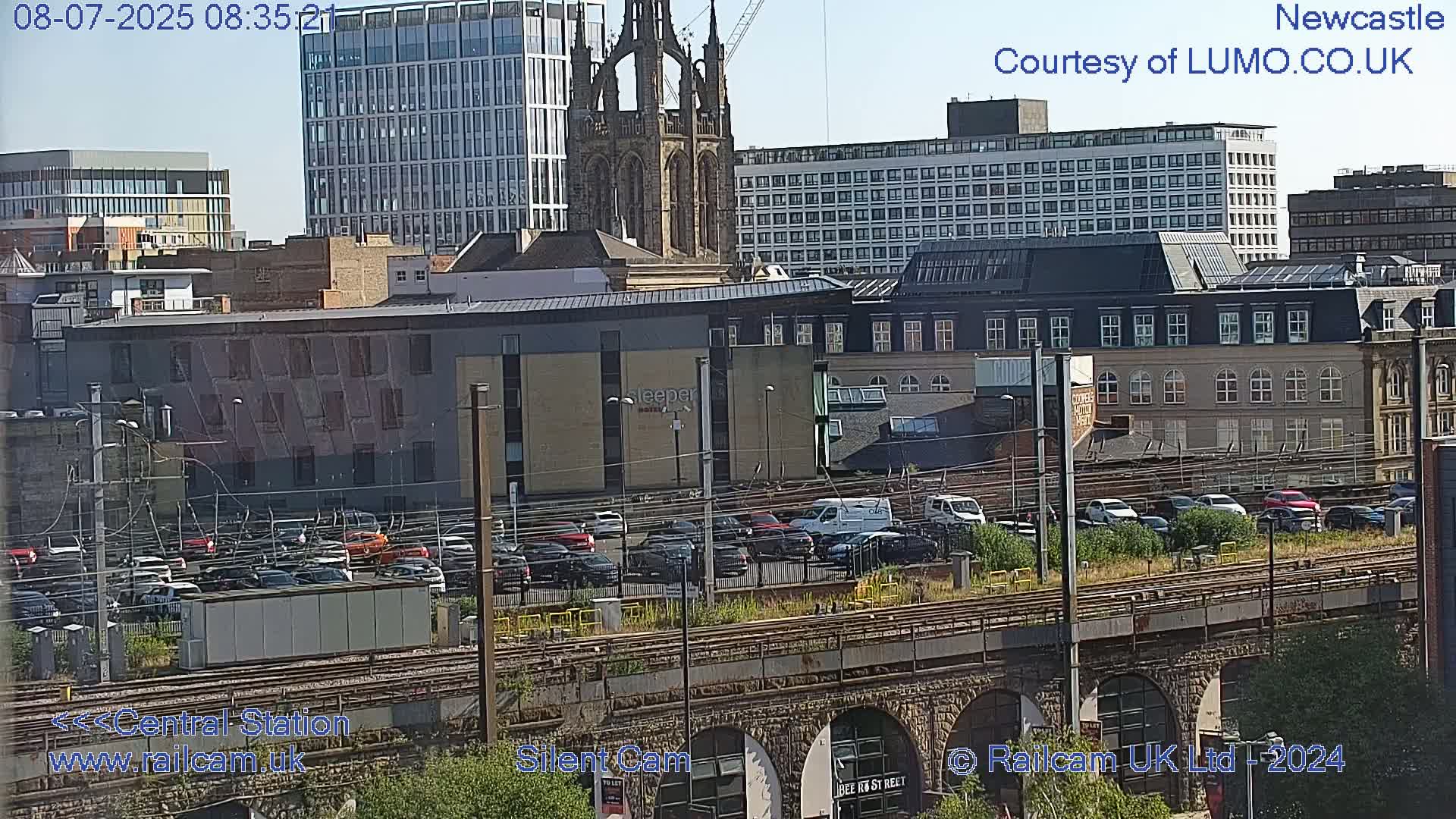 A sunny cityscape view of a city's railway tracks and buildings, including a large modern building and an older church.