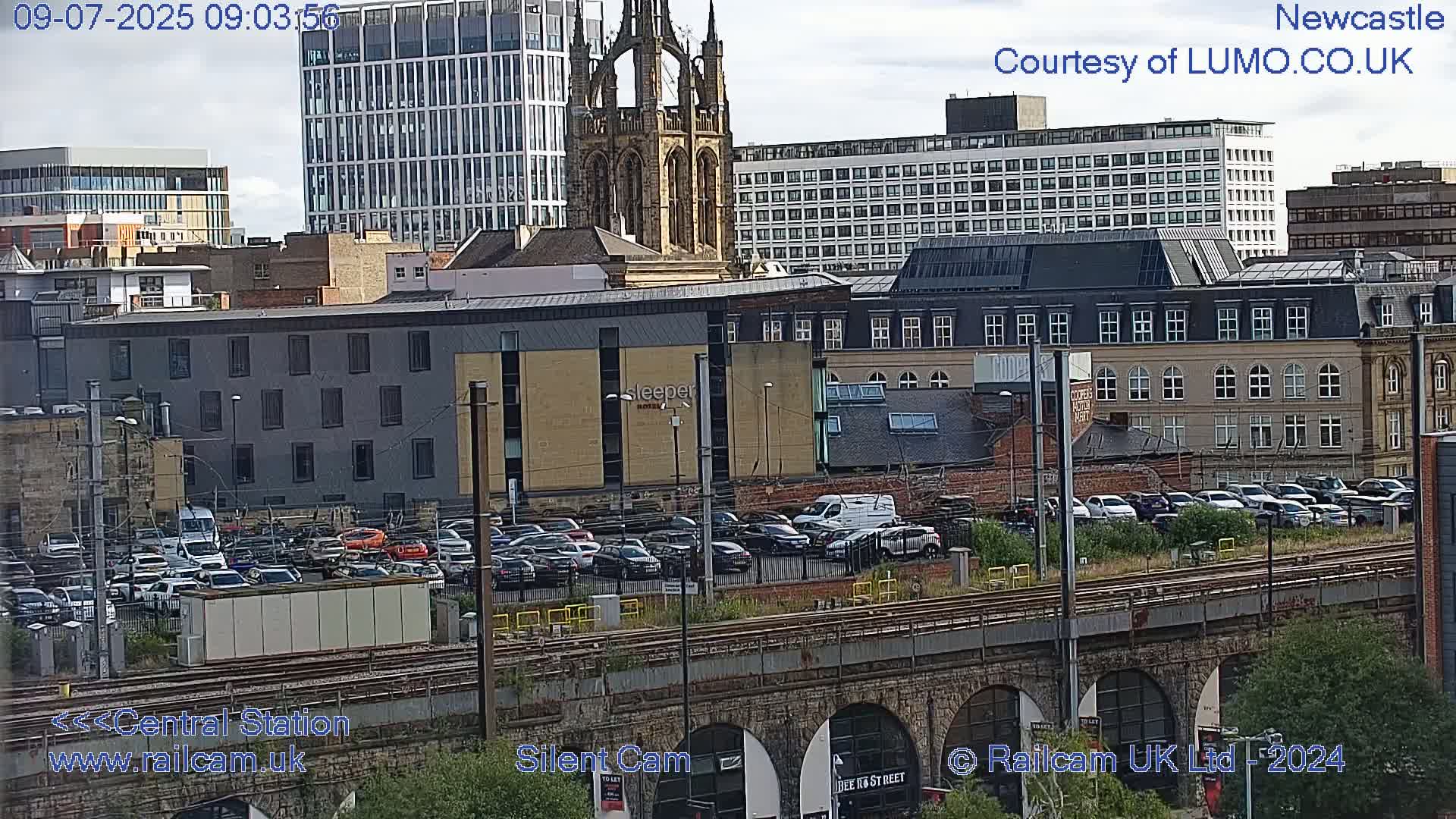 A city skyline with several buildings, a large parking lot filled with cars, and train tracks under a partly cloudy sky.