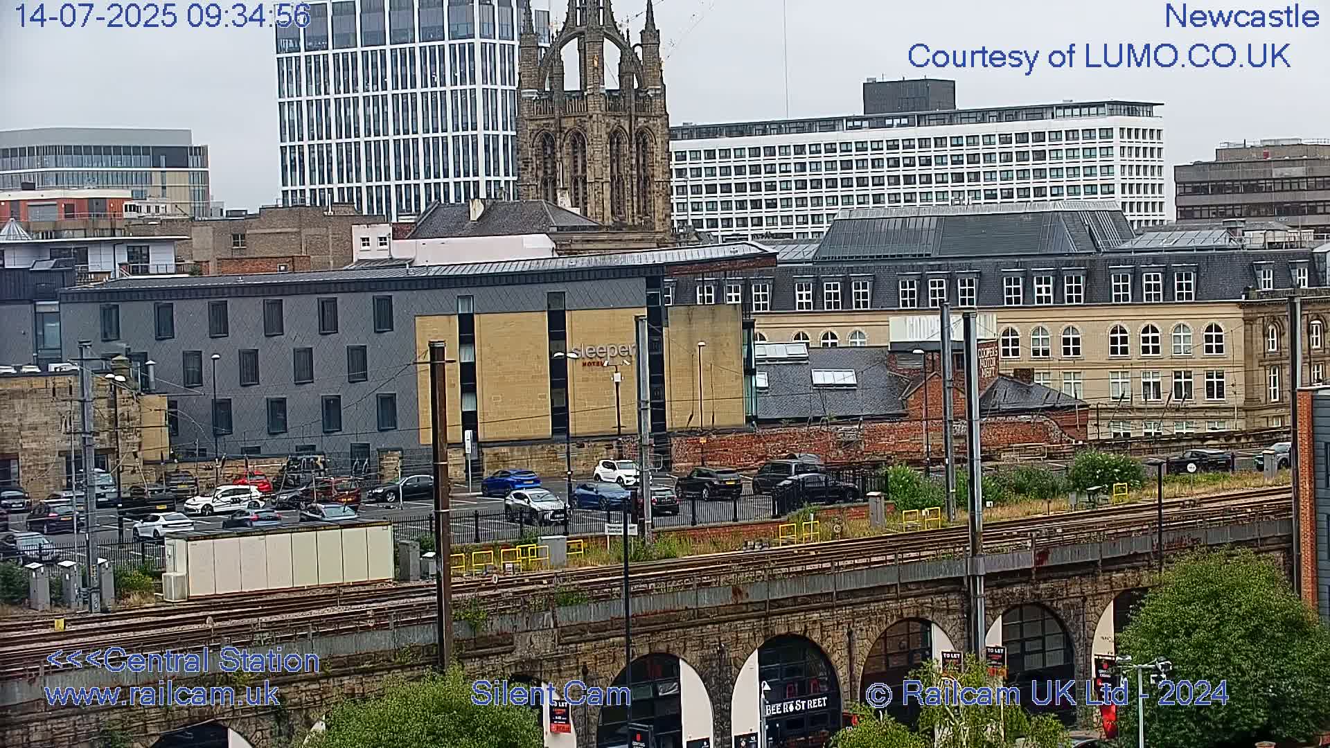 A city scene on an overcast day, showing a mix of modern and older buildings, a parking lot with cars, train tracks, and a stone railway bridge.