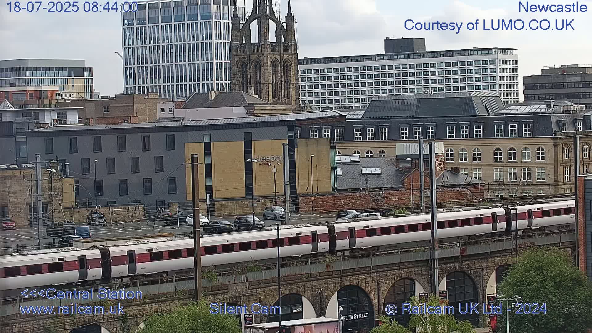 A long white and maroon train travels across a bridge over a city under an overcast sky.