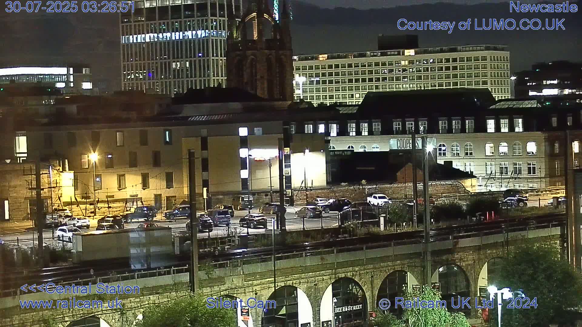 A long white and maroon train travels across a bridge over a city under an overcast sky.