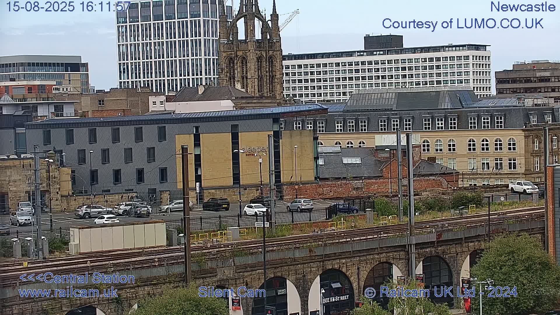 A long white and maroon train travels across a bridge over a city under an overcast sky.