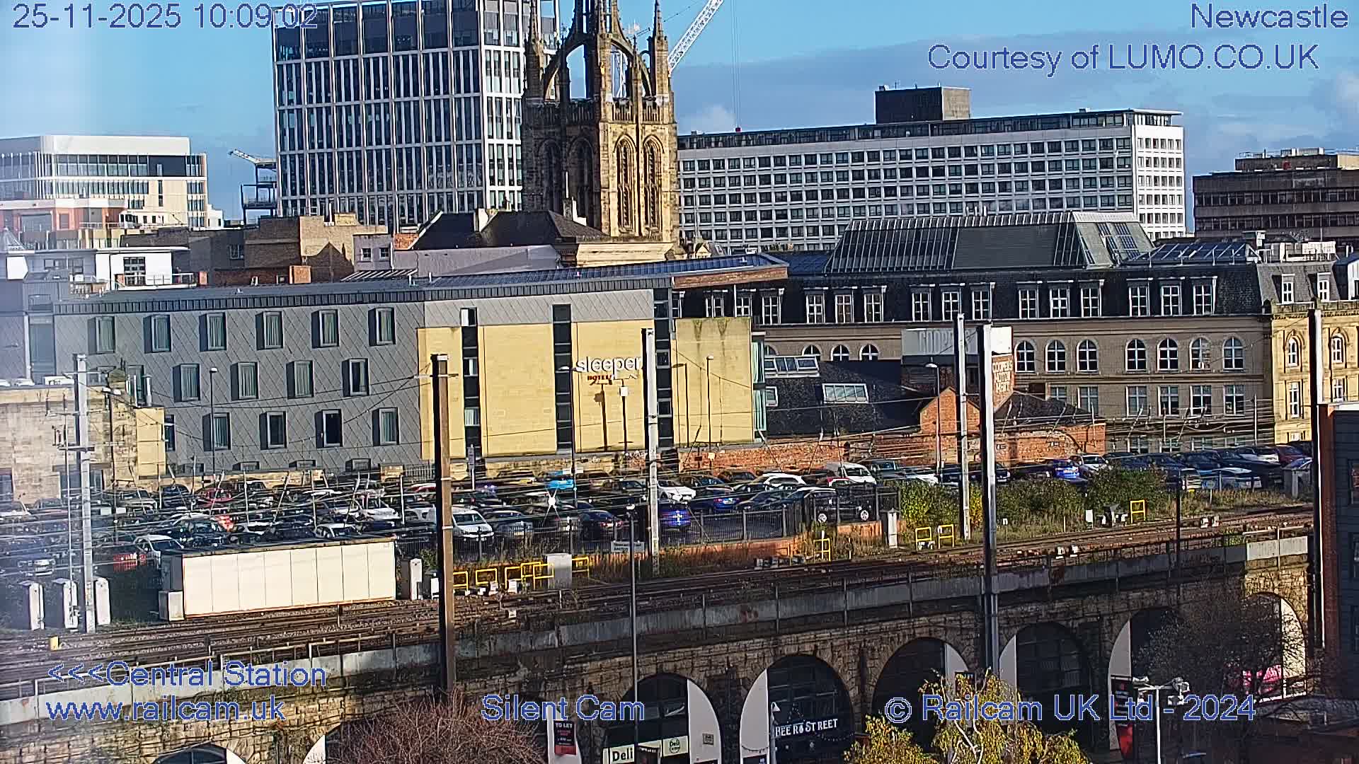 A daytime cityscape reveals a stone railway viaduct and tracks in the foreground, leading to a large car park and diverse buildings, including a prominent church tower and modern high-rises, all set against a sky with scattered clouds.