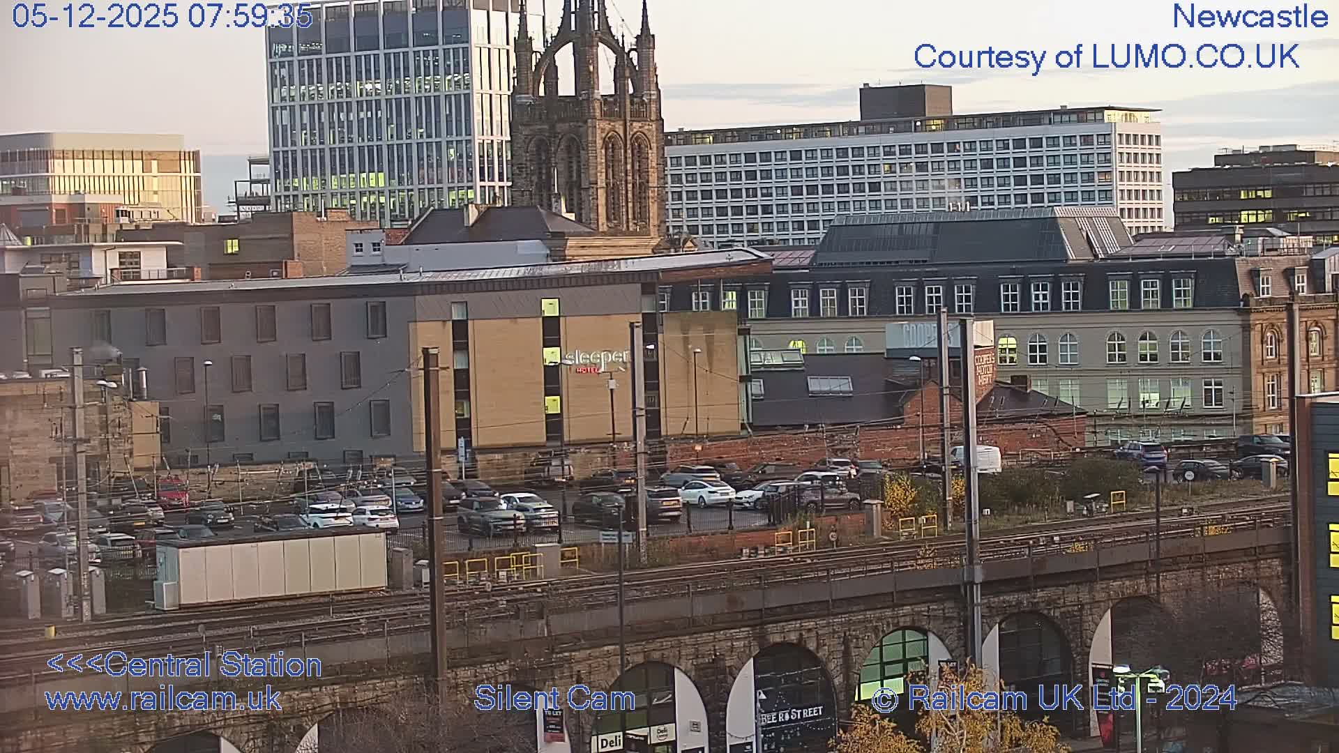 An expansive urban panorama captured on a clear morning, featuring train tracks on a stone viaduct in the foreground, a busy parking lot, and a dense arrangement of modern and historic city buildings, including a prominent church spire and glass skyscrapers, under a bright, partly cloudy sky.
