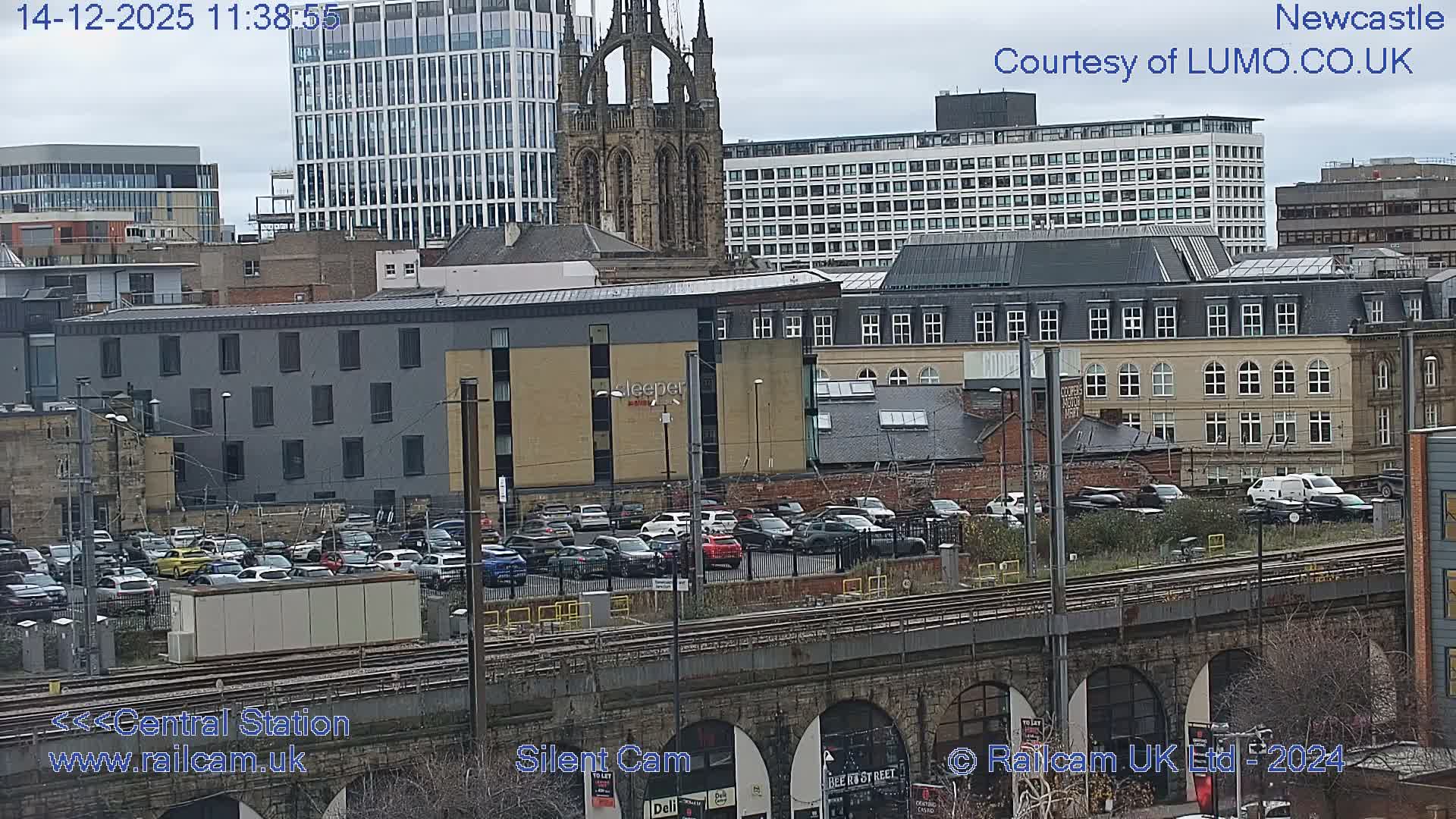 An expansive urban panorama captured on a clear morning, featuring train tracks on a stone viaduct in the foreground, a busy parking lot, and a dense arrangement of modern and historic city buildings, including a prominent church spire and glass skyscrapers, under a bright, partly cloudy sky.
