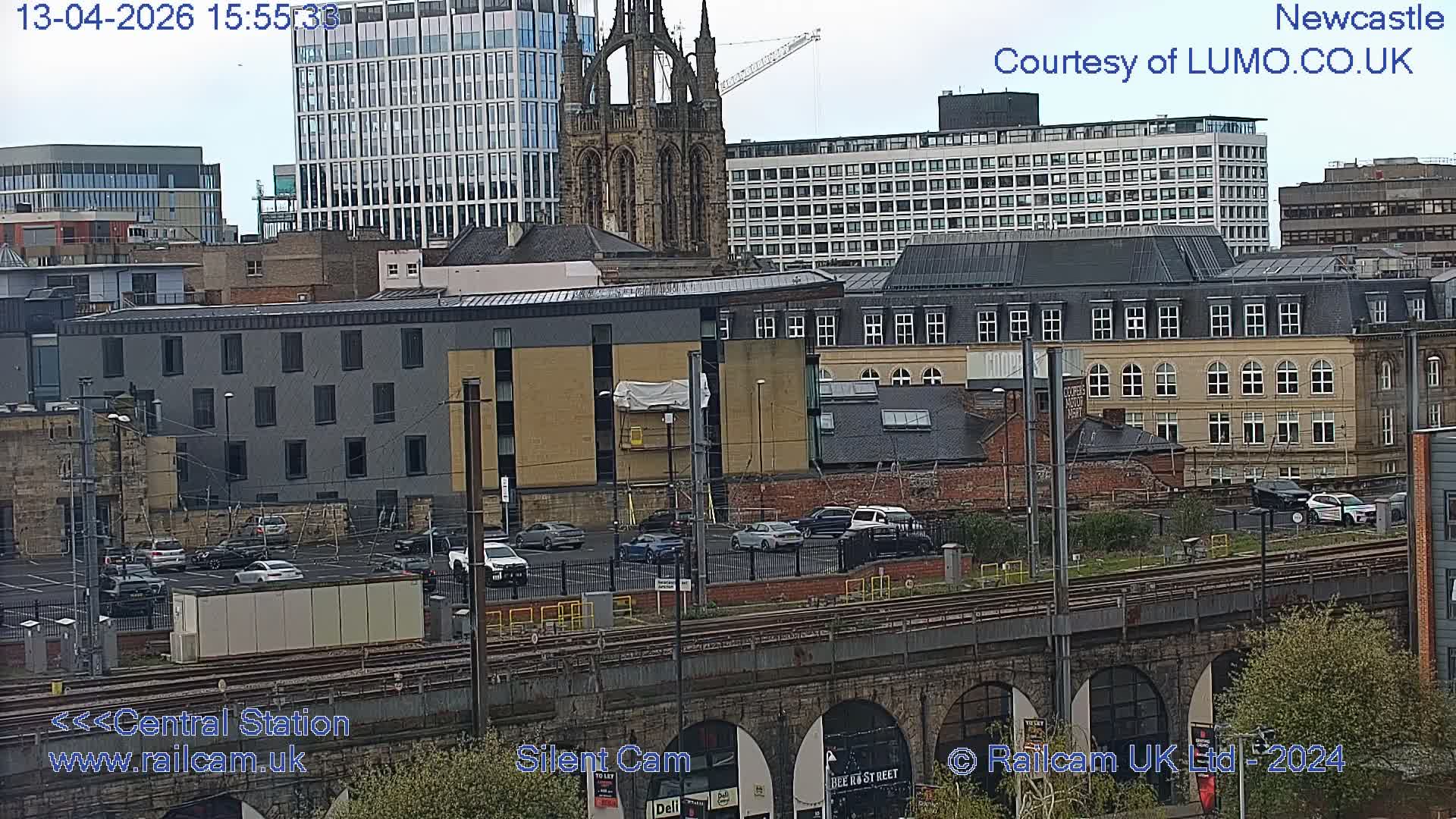 An expansive urban panorama captured on a clear morning, featuring train tracks on a stone viaduct in the foreground, a busy parking lot, and a dense arrangement of modern and historic city buildings, including a prominent church spire and glass skyscrapers, under a bright, partly cloudy sky.