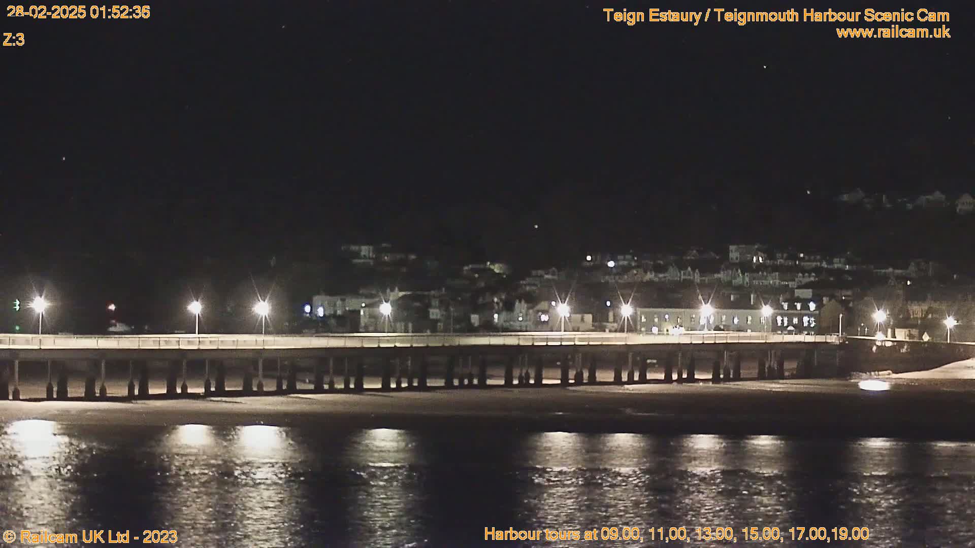 A long pier is illuminated at night by lights along its length, overlooking a calm body of water and a town in the distance under a clear, dark sky.