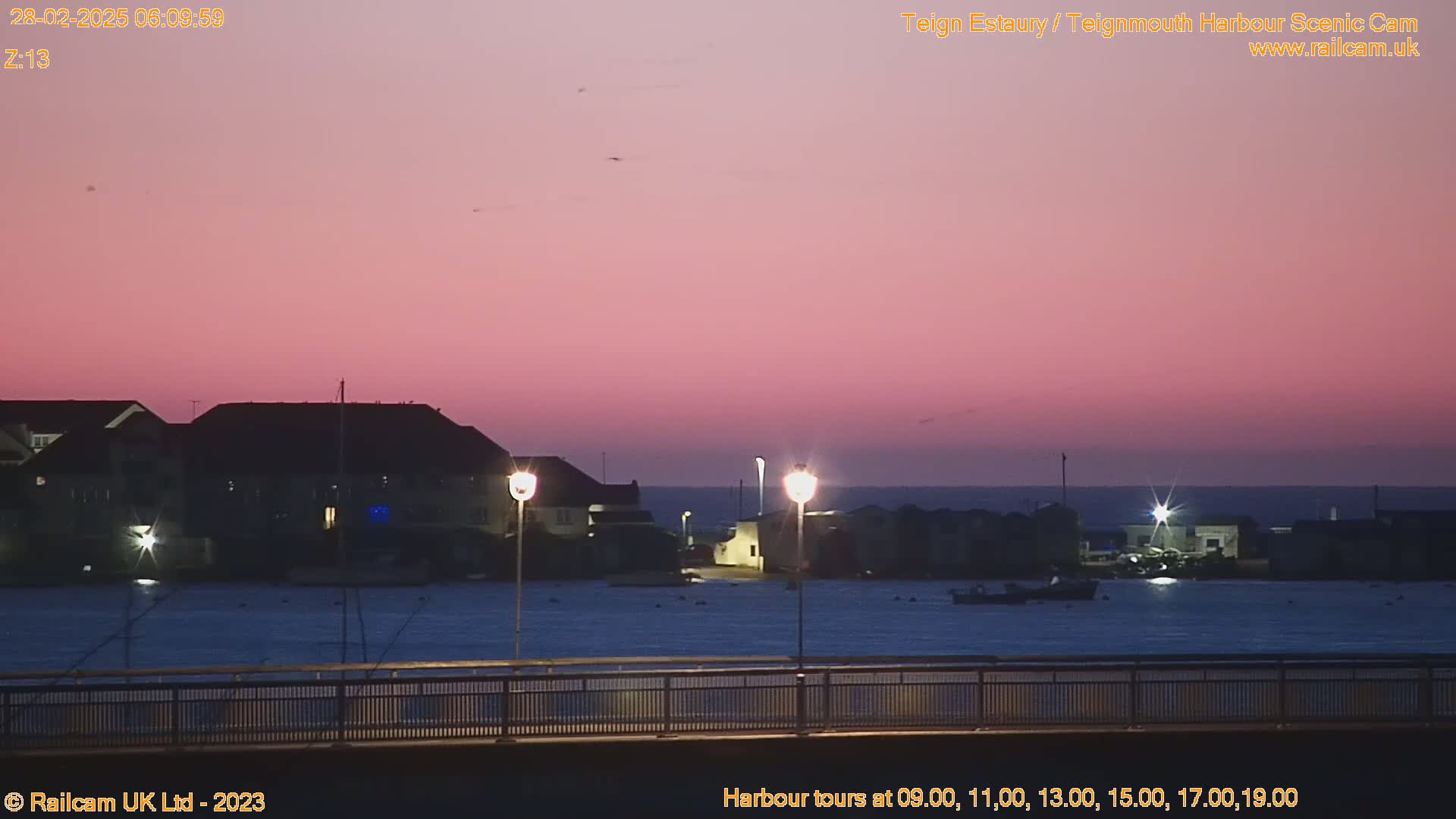 A pink-hued sunrise illuminates a calm harbor, where buildings and lights are visible along the waterfront.