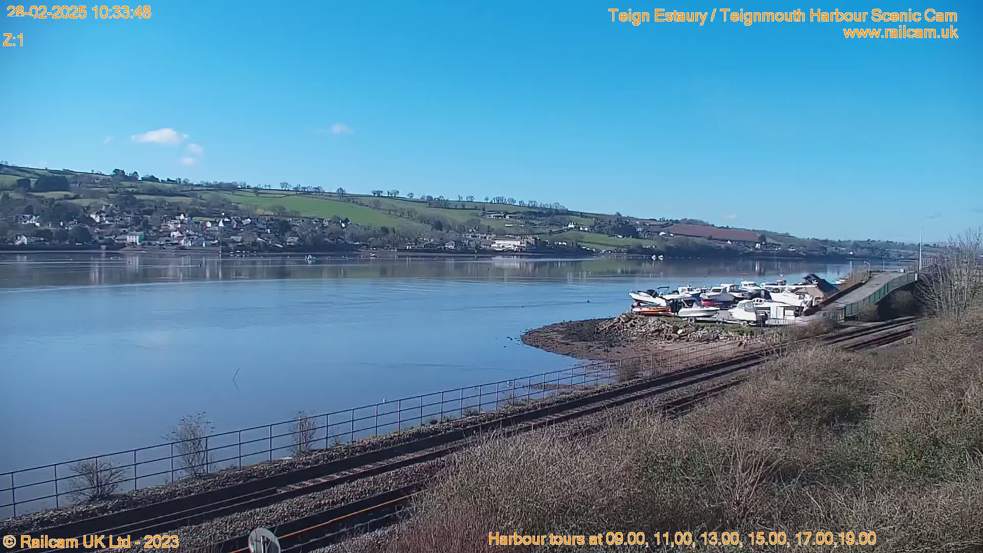 A calm, sunny day overlooks a river with docked boats, a small town on the far bank, and train tracks in the foreground.