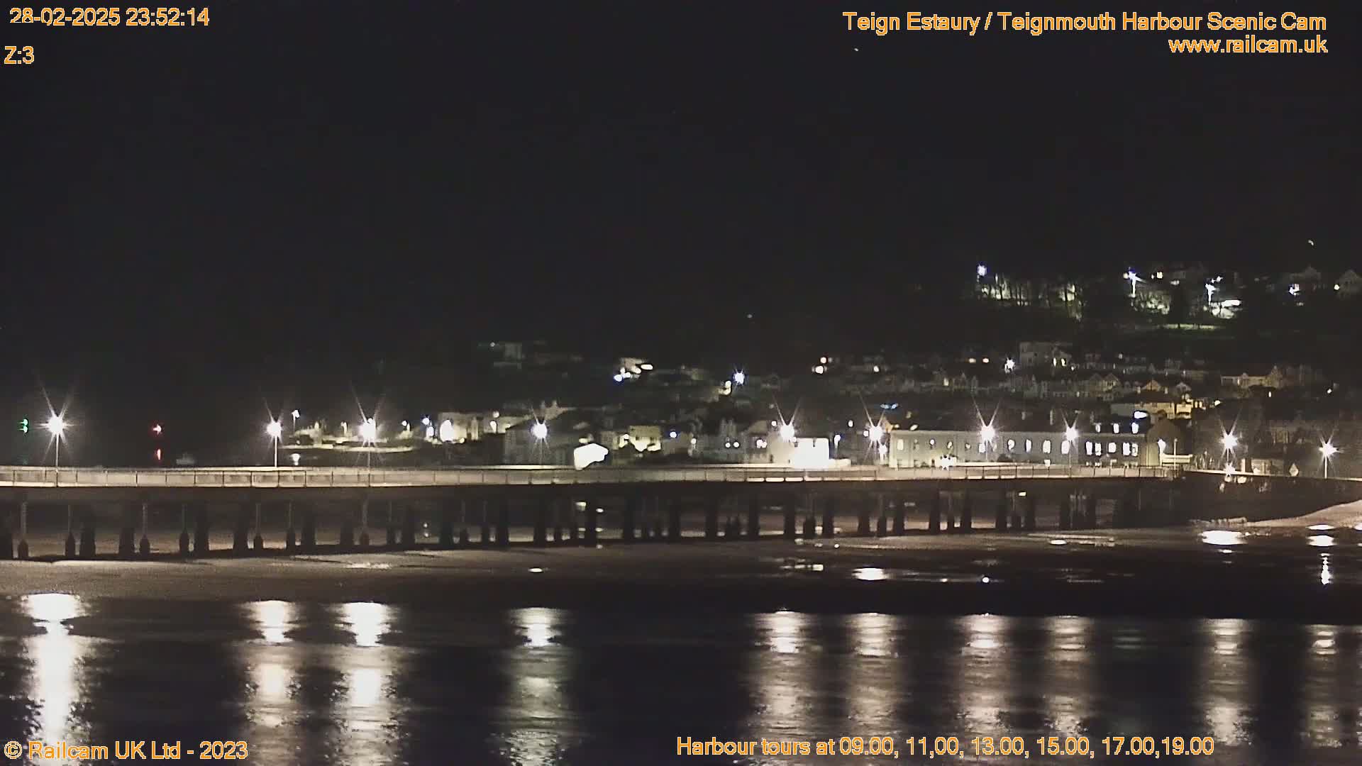 A long pier stretches out over a dark, calm body of water at night, with a town's lights visible in the distance.