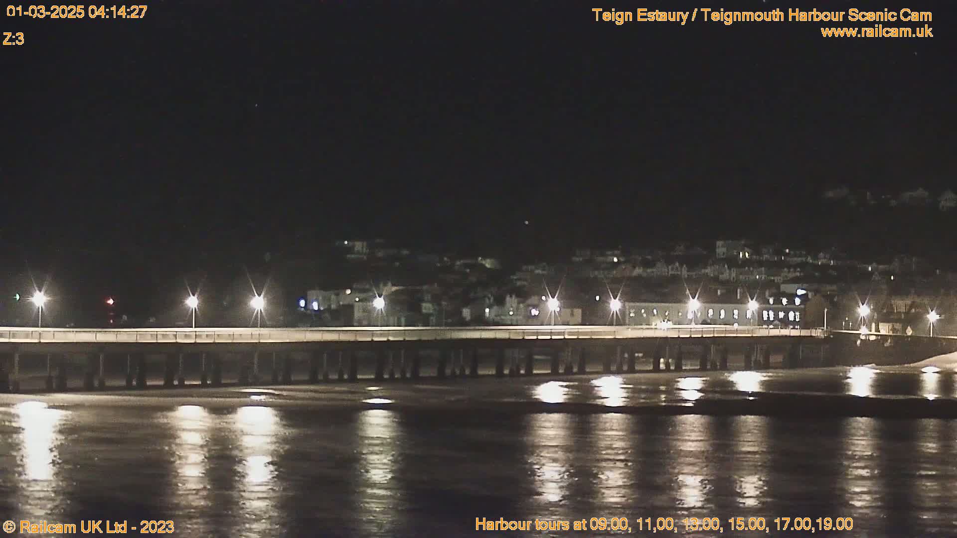 A long pier is illuminated at night, extending over dark, calm water towards a dimly lit town.