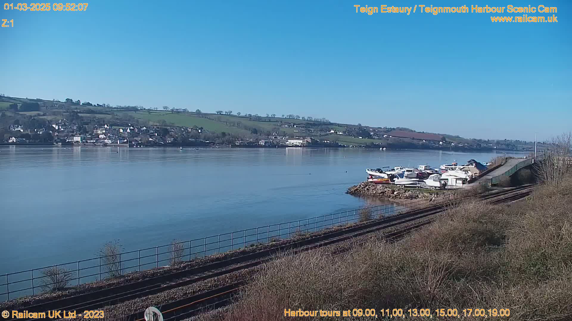 A calm river with several boats docked near a railway line under a clear blue sky.