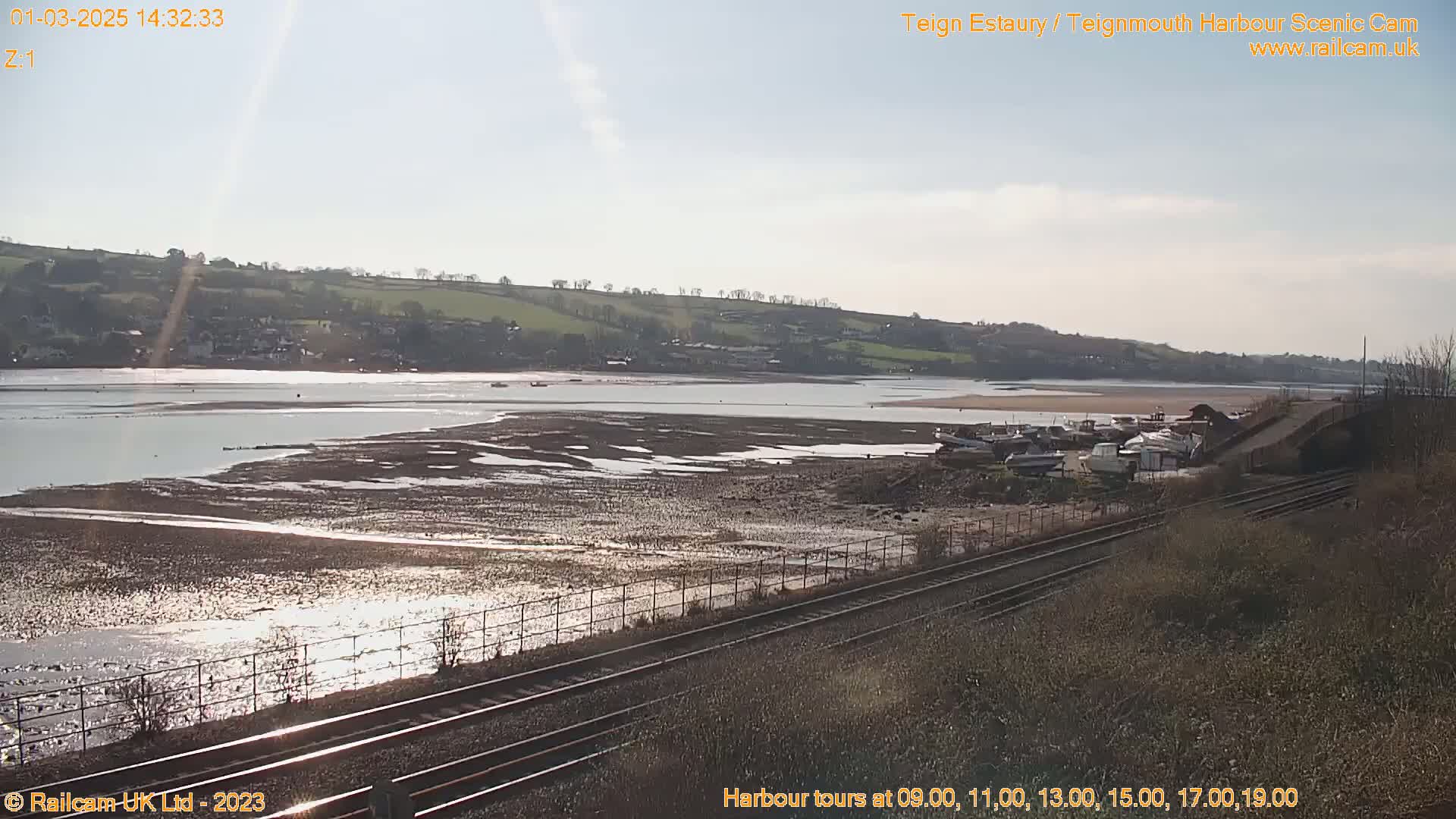 A wide shot of a harbor at low tide on a sunny day shows several boats resting on the exposed mudflats, with a railway line in the foreground.