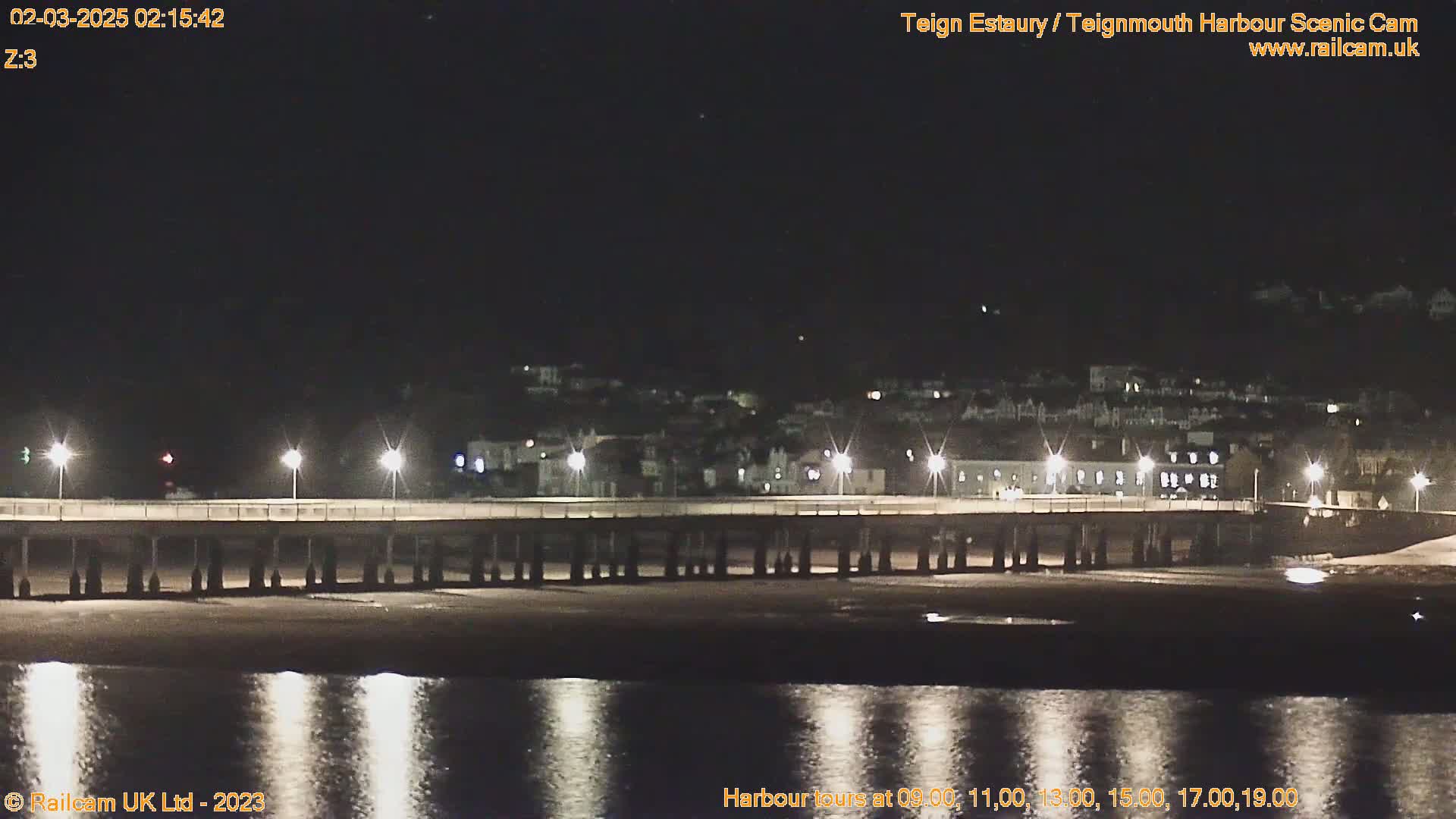 A long pier at night, illuminated by lights, extends over calm water reflecting the lights, with a town visible in the background under a clear, dark sky.