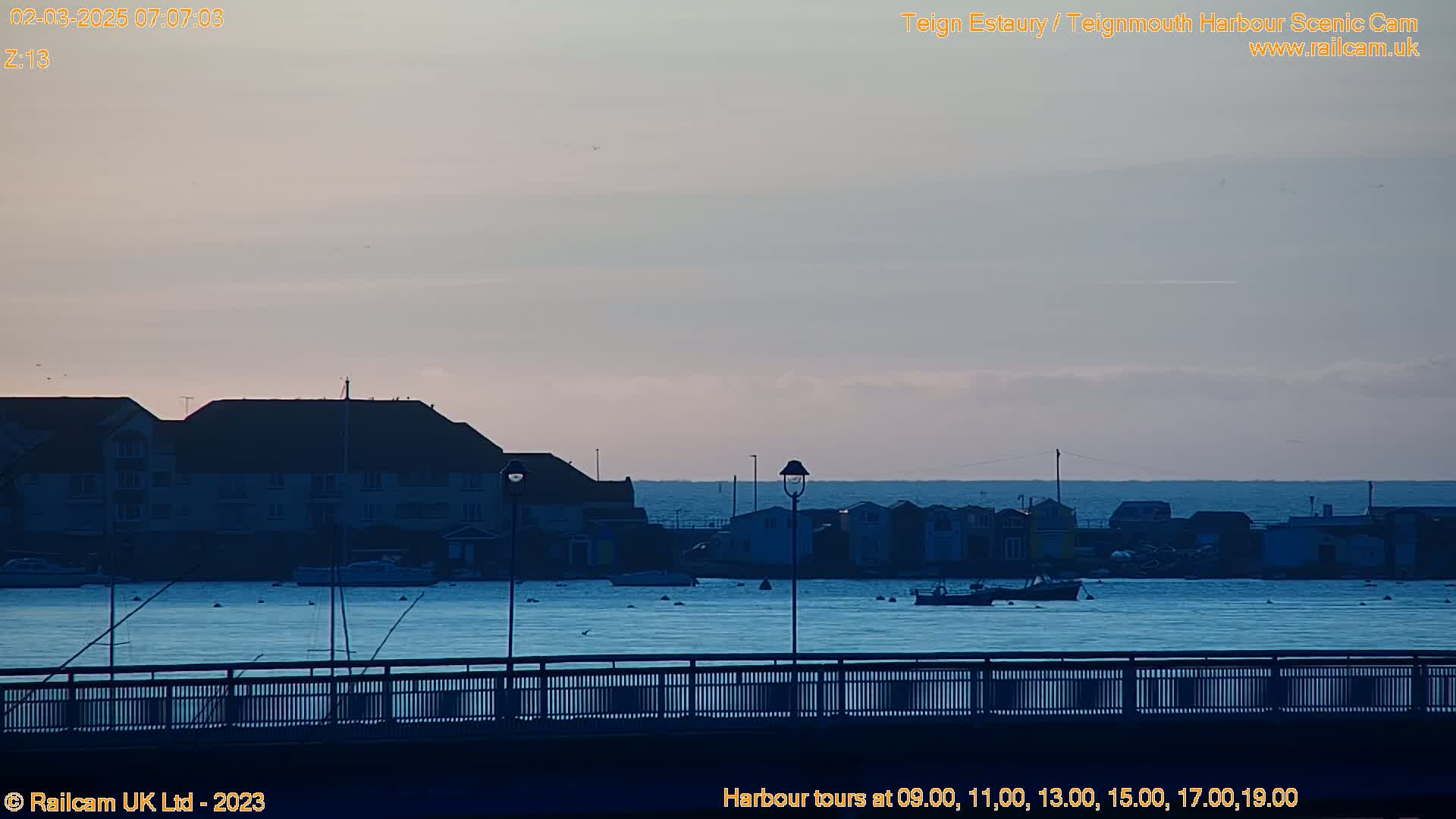 A calm harbor scene at dawn, with buildings and boats visible across the water, viewed from a bridge.