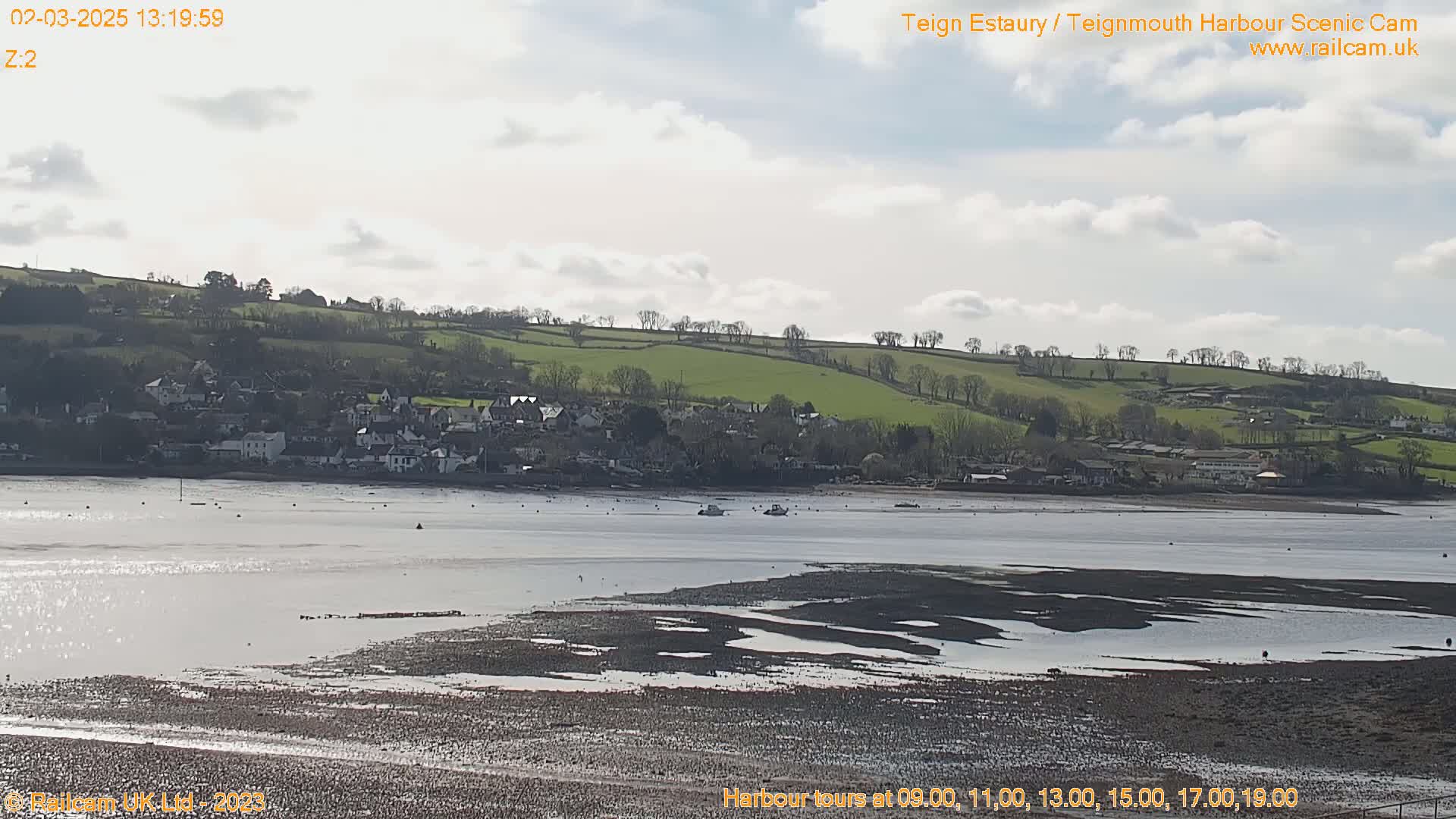 A mostly sunny day reveals a calm estuary with exposed mudflats in the foreground, a small town nestled against a gently sloping green hill in the background.