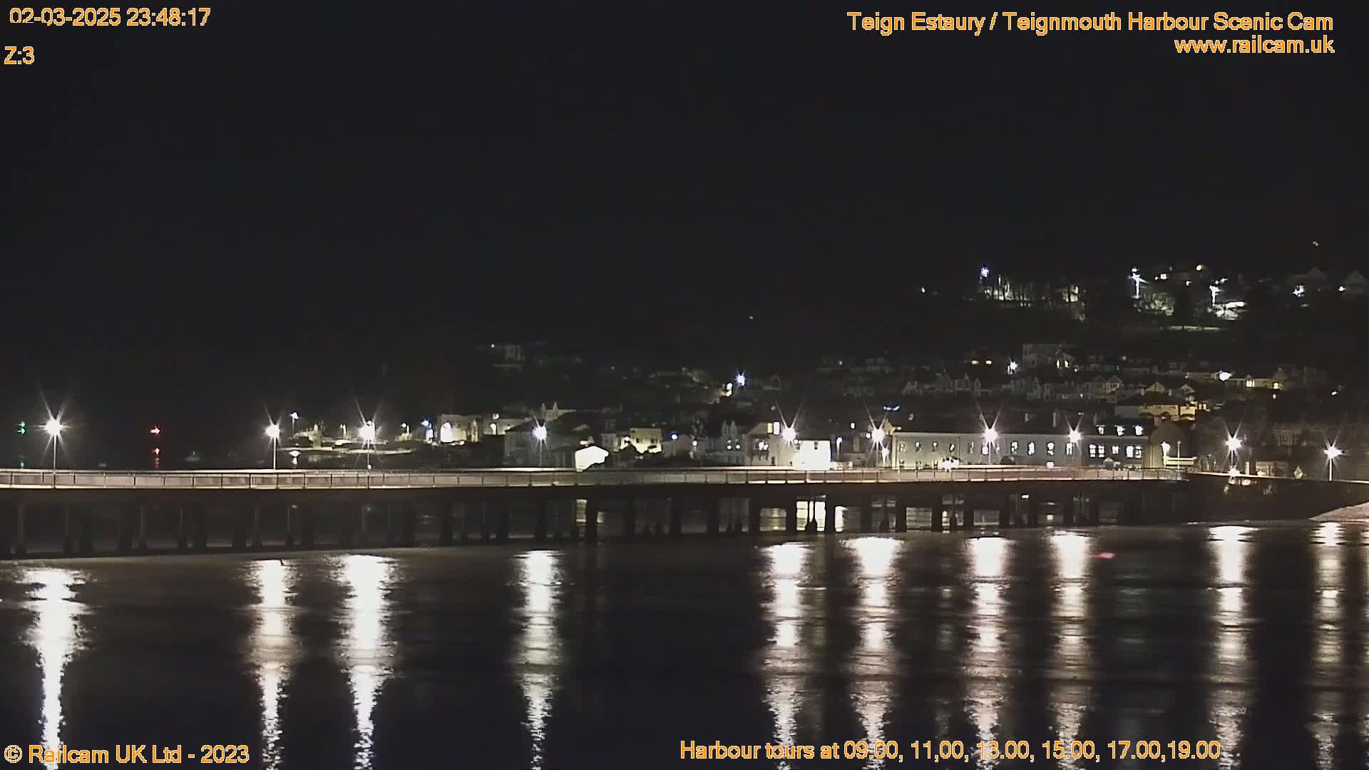 A long pier stretches across a dark, calm body of water reflecting the lights of a town on the far shore at night.