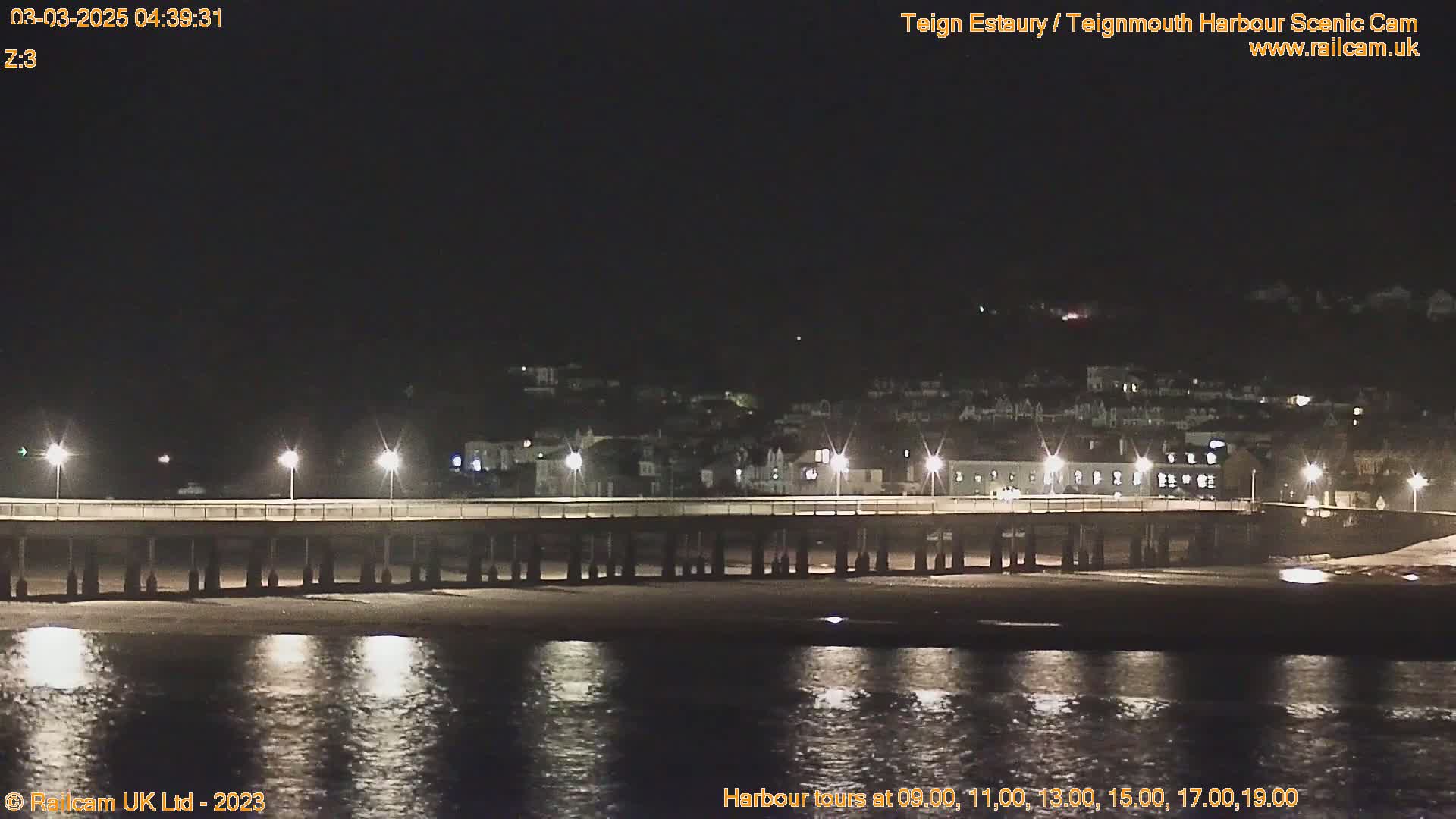 A long pier stretches across a dark, calm body of water at night, with a town's lights visible in the background.
