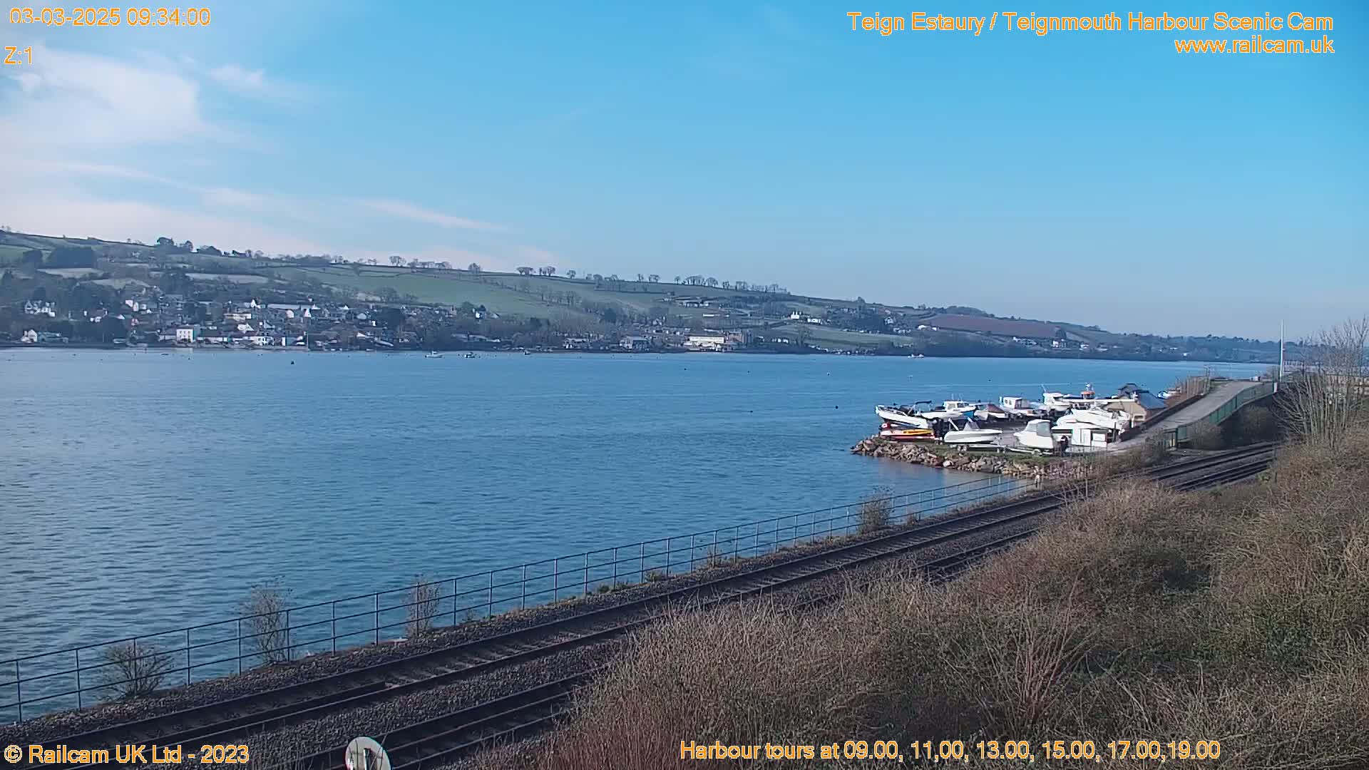 A calm body of water, under a clear blue sky, shows several boats docked near a railway track and a small town on the far shore.