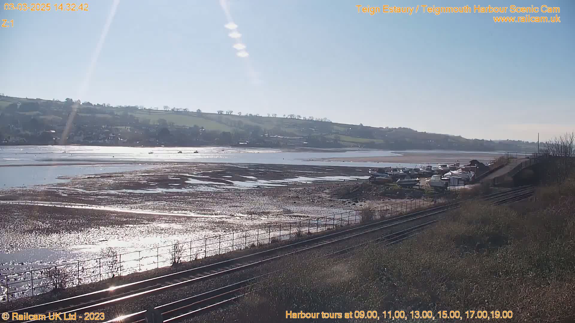 A sunny day reveals a wide estuary at low tide, with boats beached near a railway line and hills in the background.