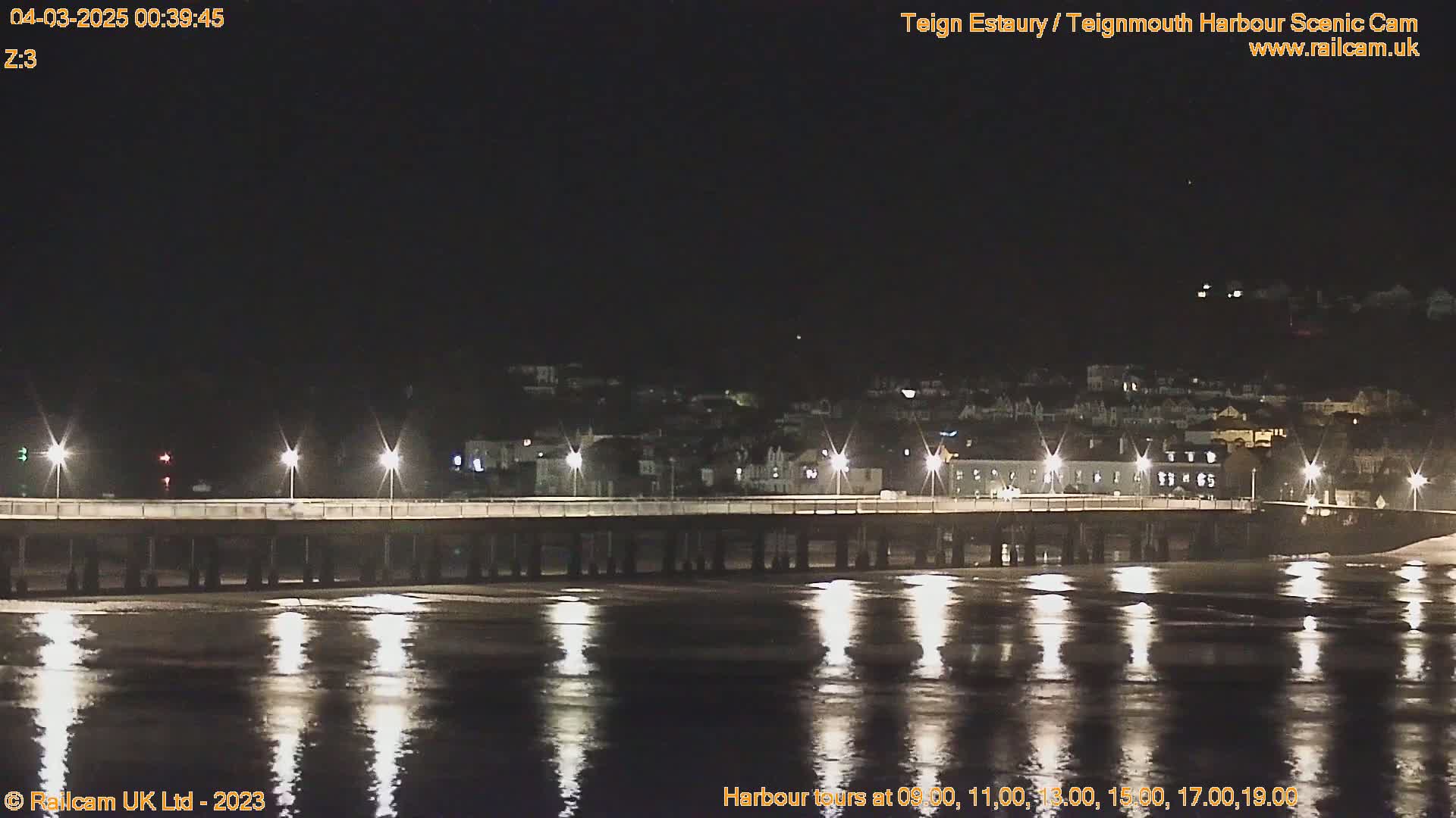 A long pier stretches across the water at night, lit by several lights, with a town visible in the distance under a clear, dark sky.