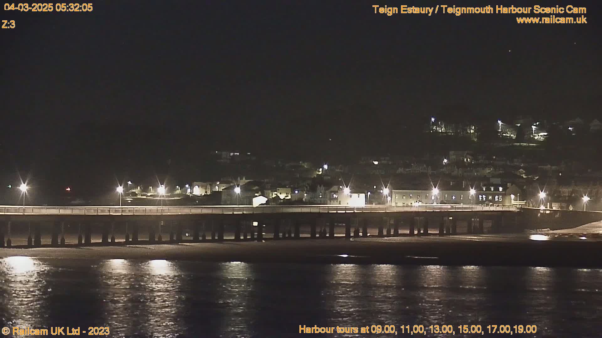 A long pier stretches across a dark, calm body of water at night, with a town illuminated by lights visible in the distance.