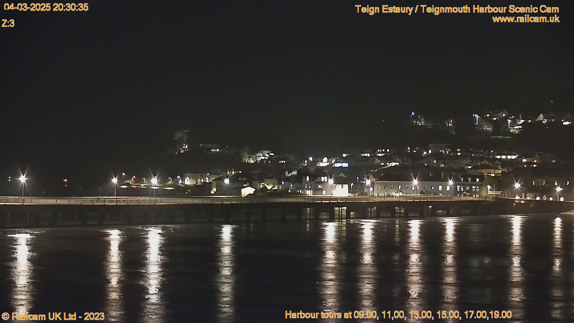 A nighttime view of a town's lights reflected in calm water, with a bridge stretching across the foreground.