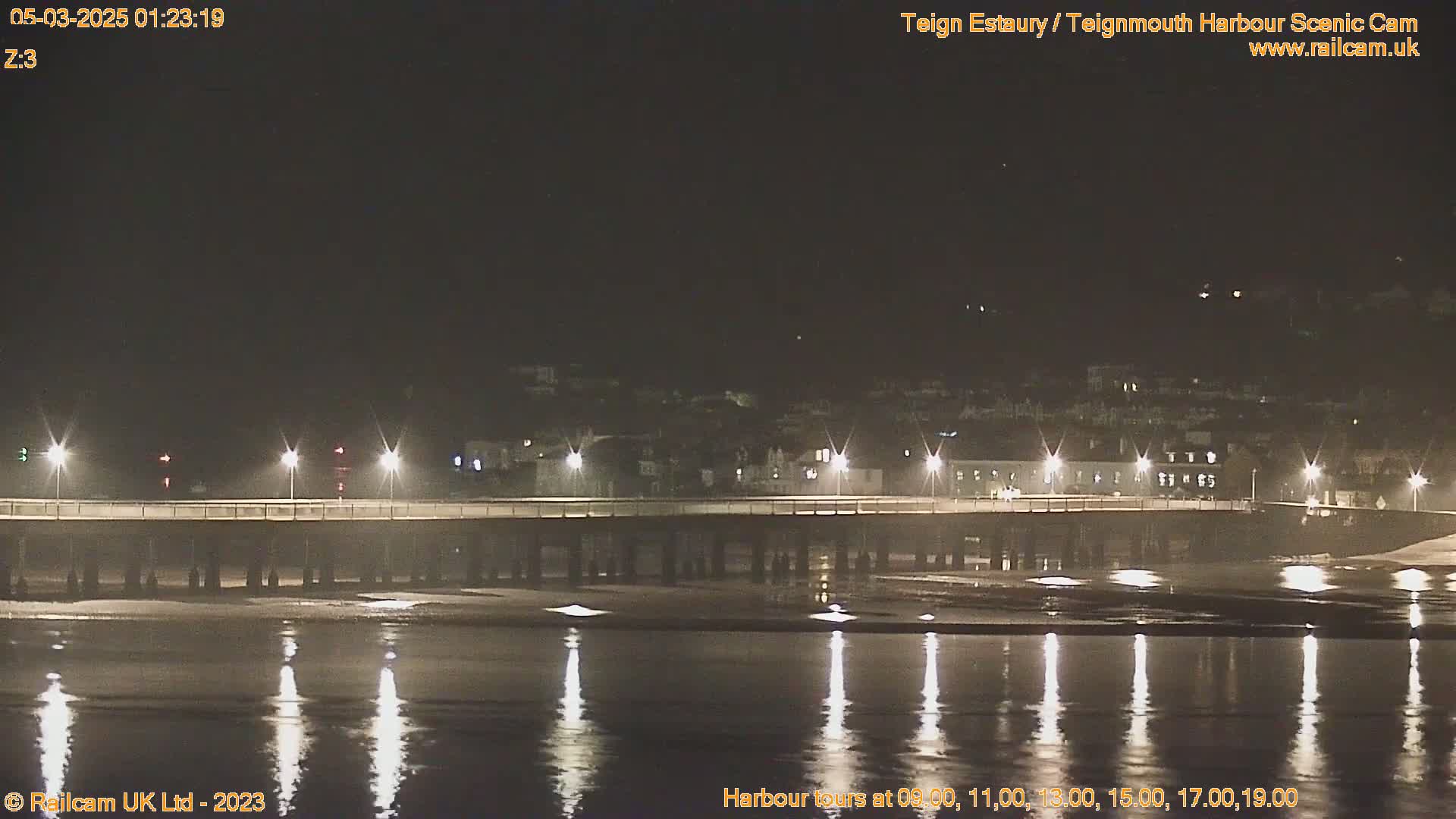 A long pier lit by streetlights stretches across a calm, dark body of water at night, reflecting the lights onto the water's surface.