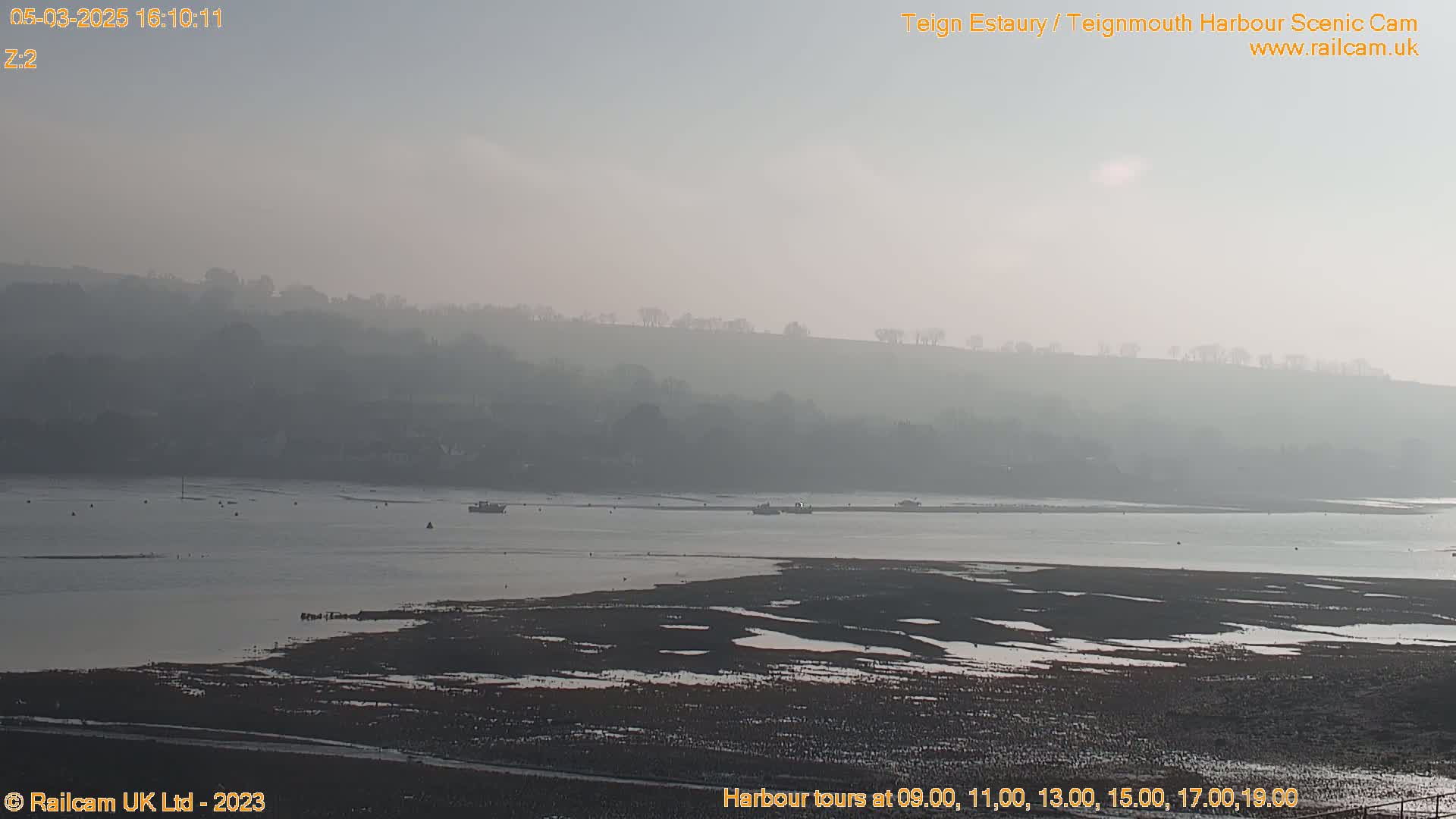 A hazy, calm day reveals a wide estuary with several small boats and a flat, mud-covered shoreline under a pale sky.