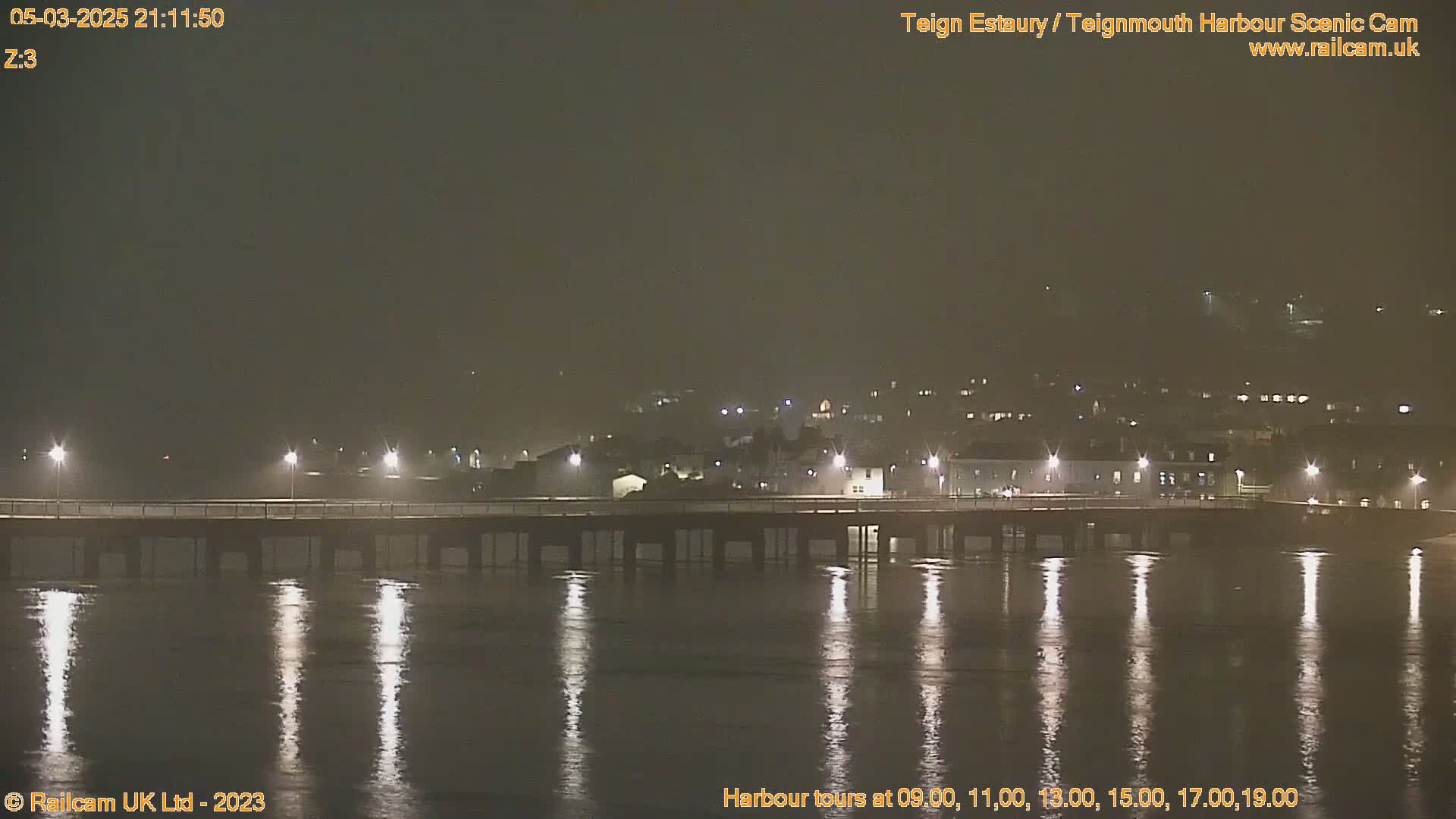 A long pier is illuminated at night, extending over dark, calm water reflecting the lights, with a town visible in the distance under a dark sky.