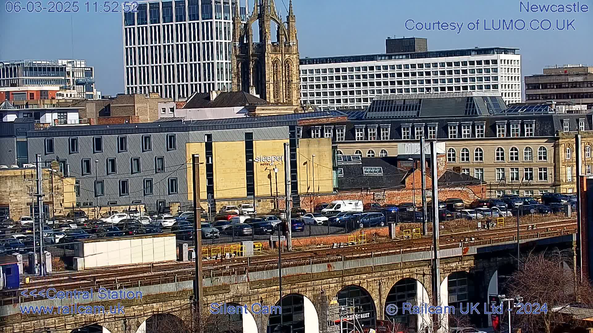 A sunny view of a city, featuring a railway bridge, train tracks, numerous parked cars, several buildings of varying architectural styles, and a church steeple in the background.
