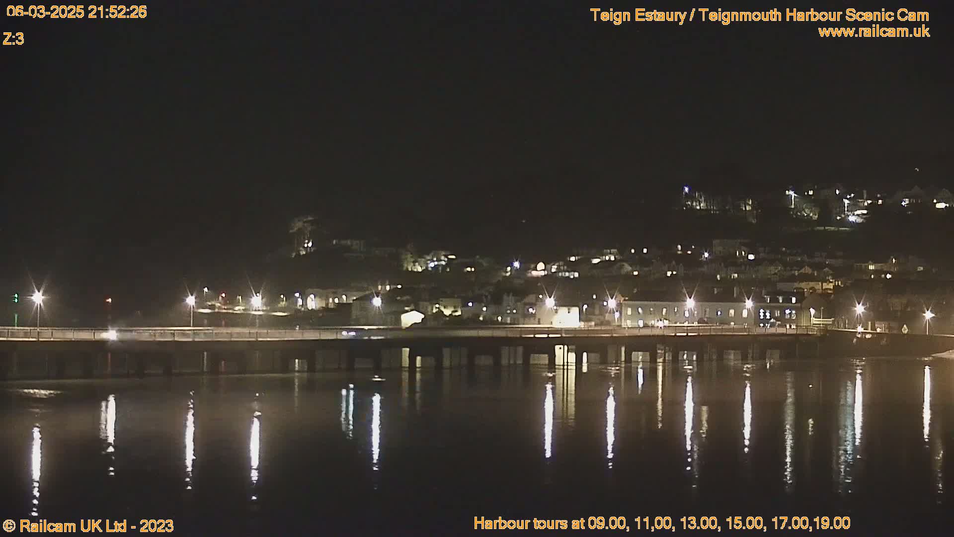 At night, a bridge illuminated by lights spans a calm body of water reflecting the lights, with a town visible on the far bank.