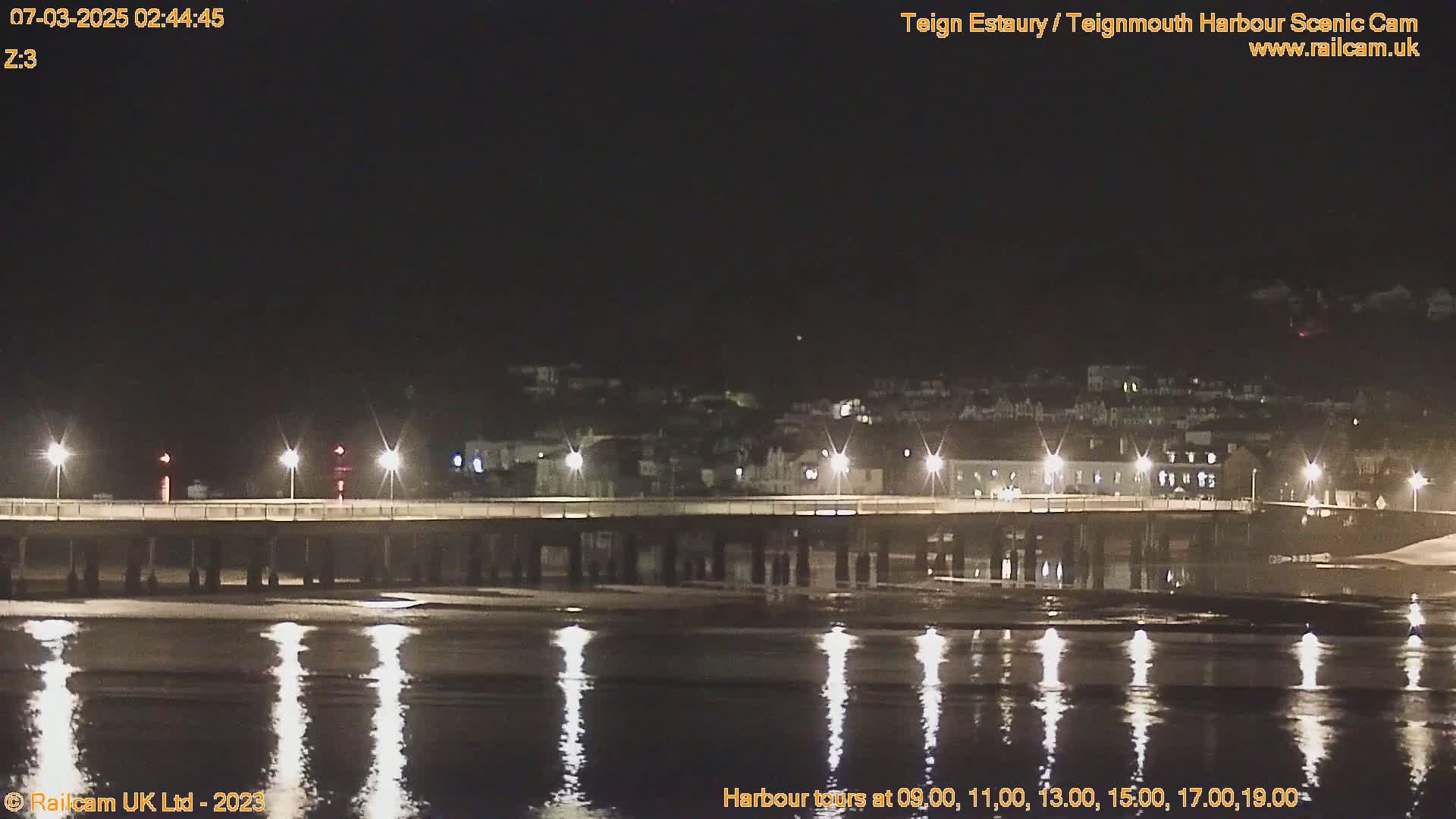 A long pier is illuminated at night, reflecting in the calm water under a dark sky.